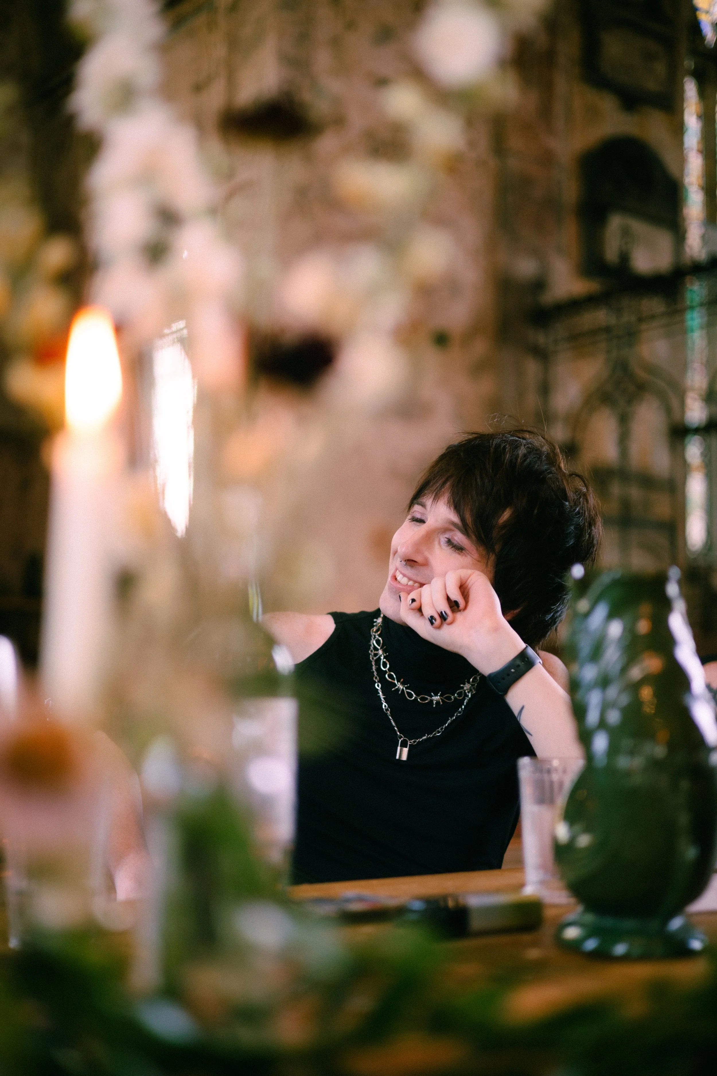 A person with short dark hair, jewelry, and makeup smiling and resting their head on their hand in a cozy, rustic indoor setting with warm lighting.