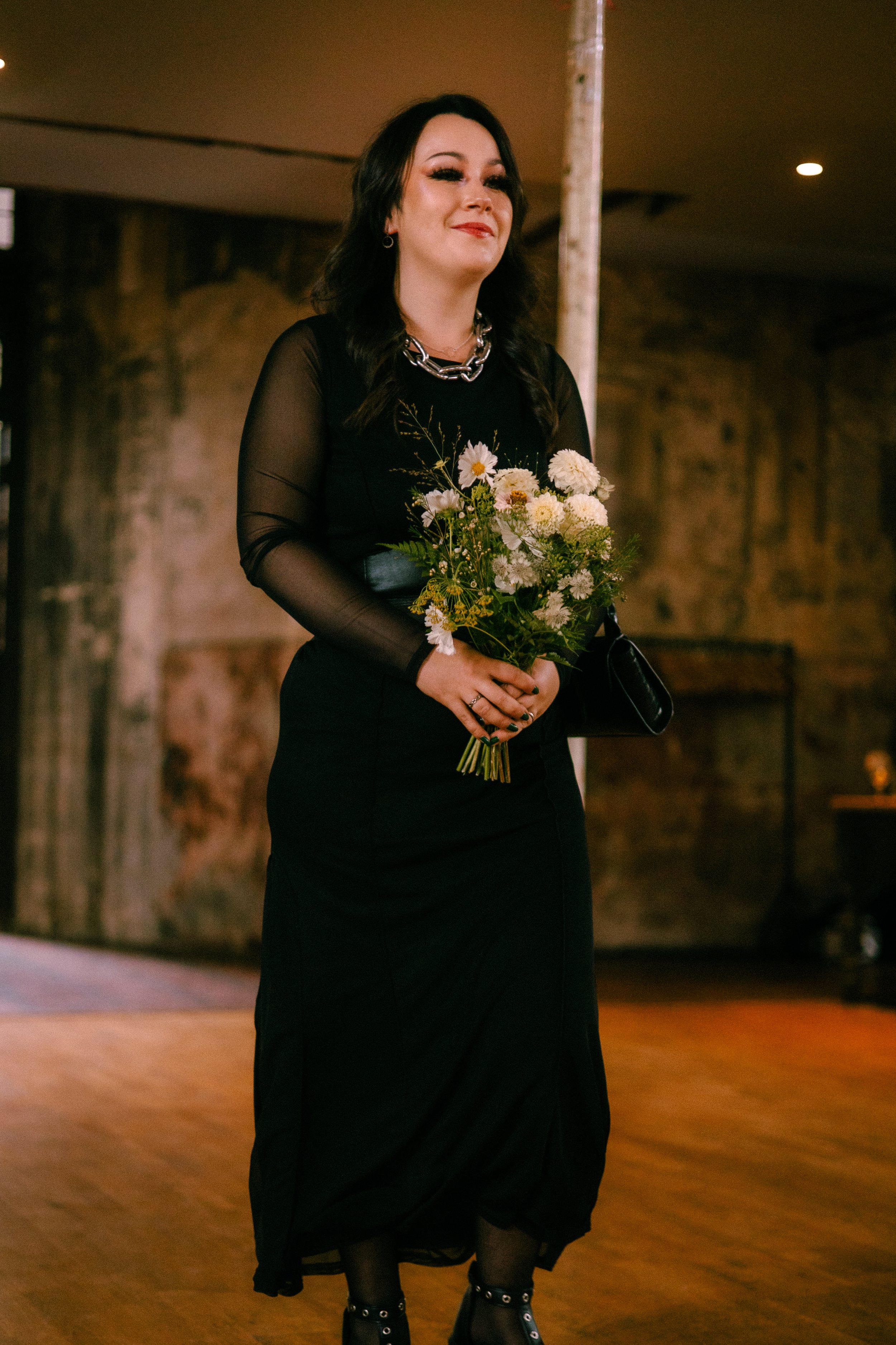 A woman with dark hair and makeup holding a bouquet of white flowers, wearing a black dress with sheer sleeves and black lace-up shoes with buckles, standing in a warmly lit indoor setting with wooden flooring and brick walls.