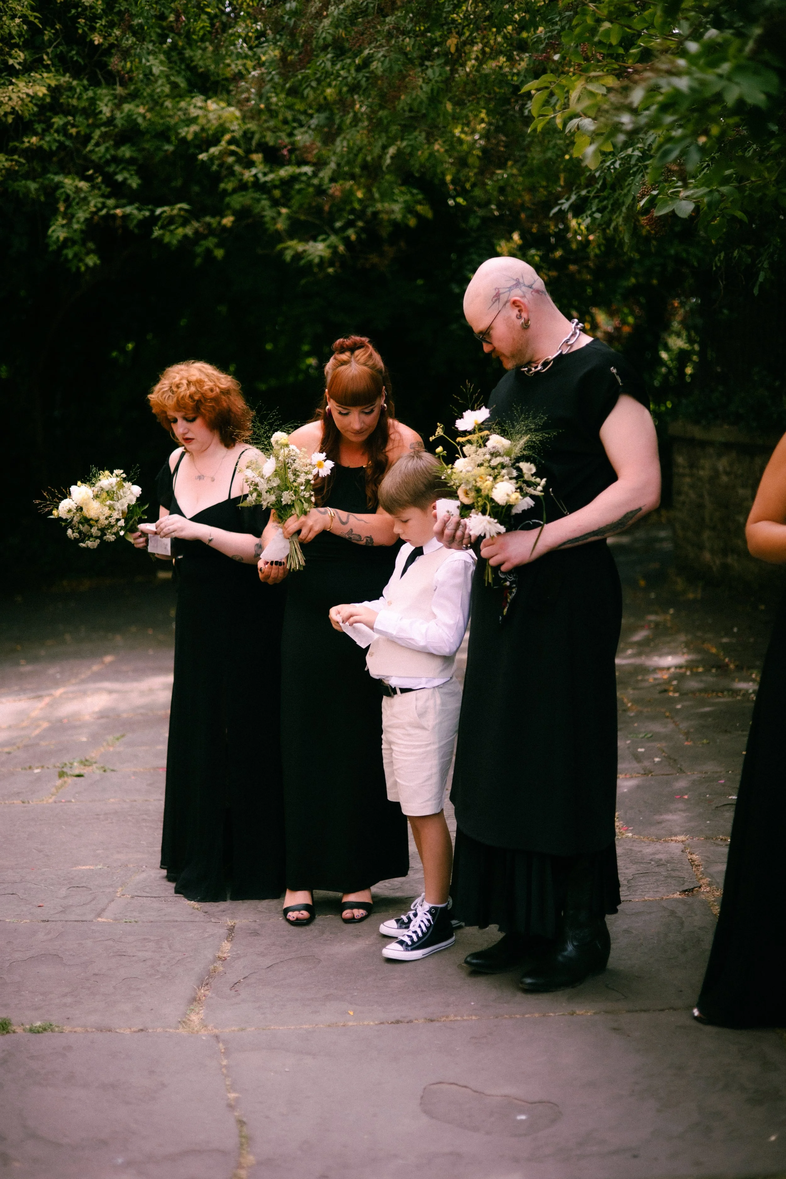 People standing outdoors holding bouquets of flowers, dressed in black, with one child in white, on a paved area surrounded by green trees.