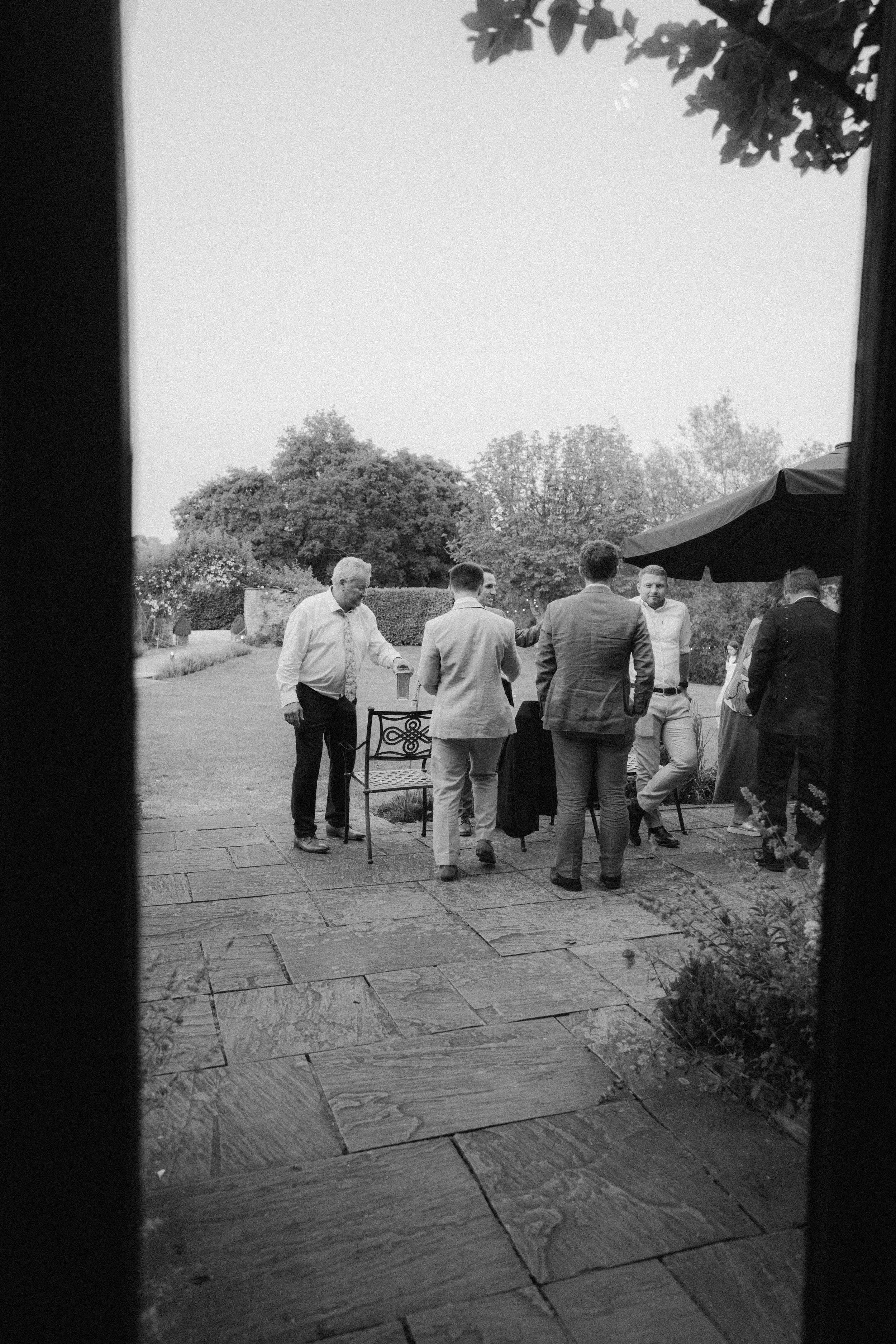 Group of men in formal attire socializing outside on a patio with trees in the background, viewed through a doorway.