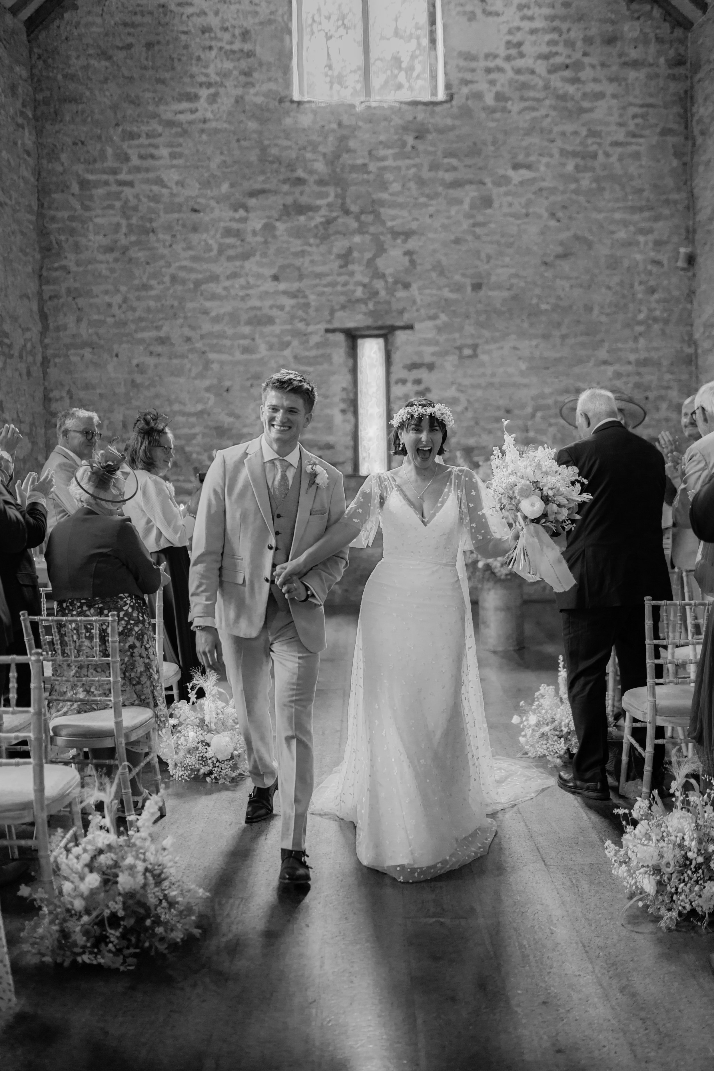 A black and white photo of a newlywed couple walking down the aisle in a rustic church, smiling and holding hands, with guests clapping on either side.