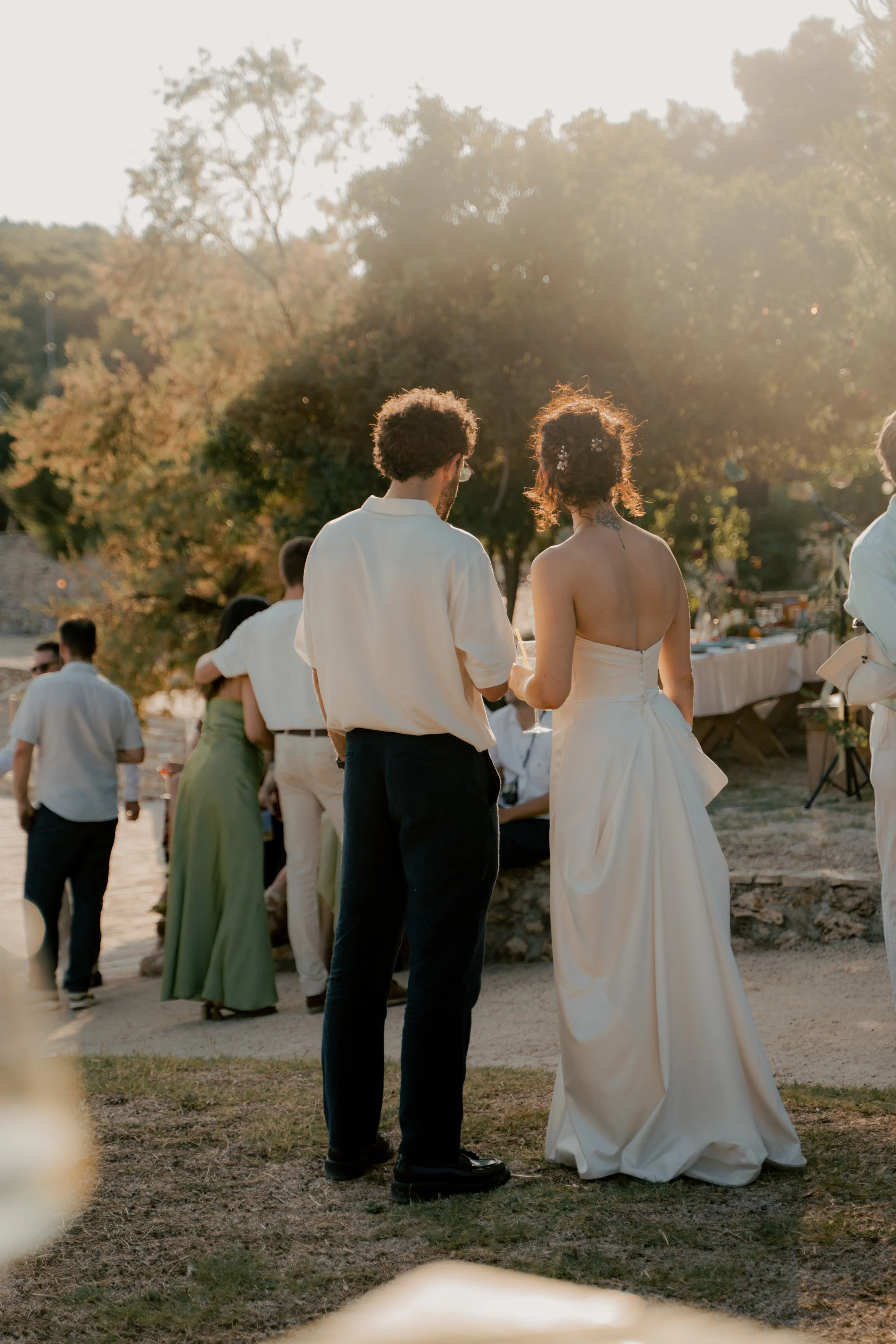 A couple dressed in formal attire at an outdoor wedding reception during sunset, with guests in the background and decorated tables.