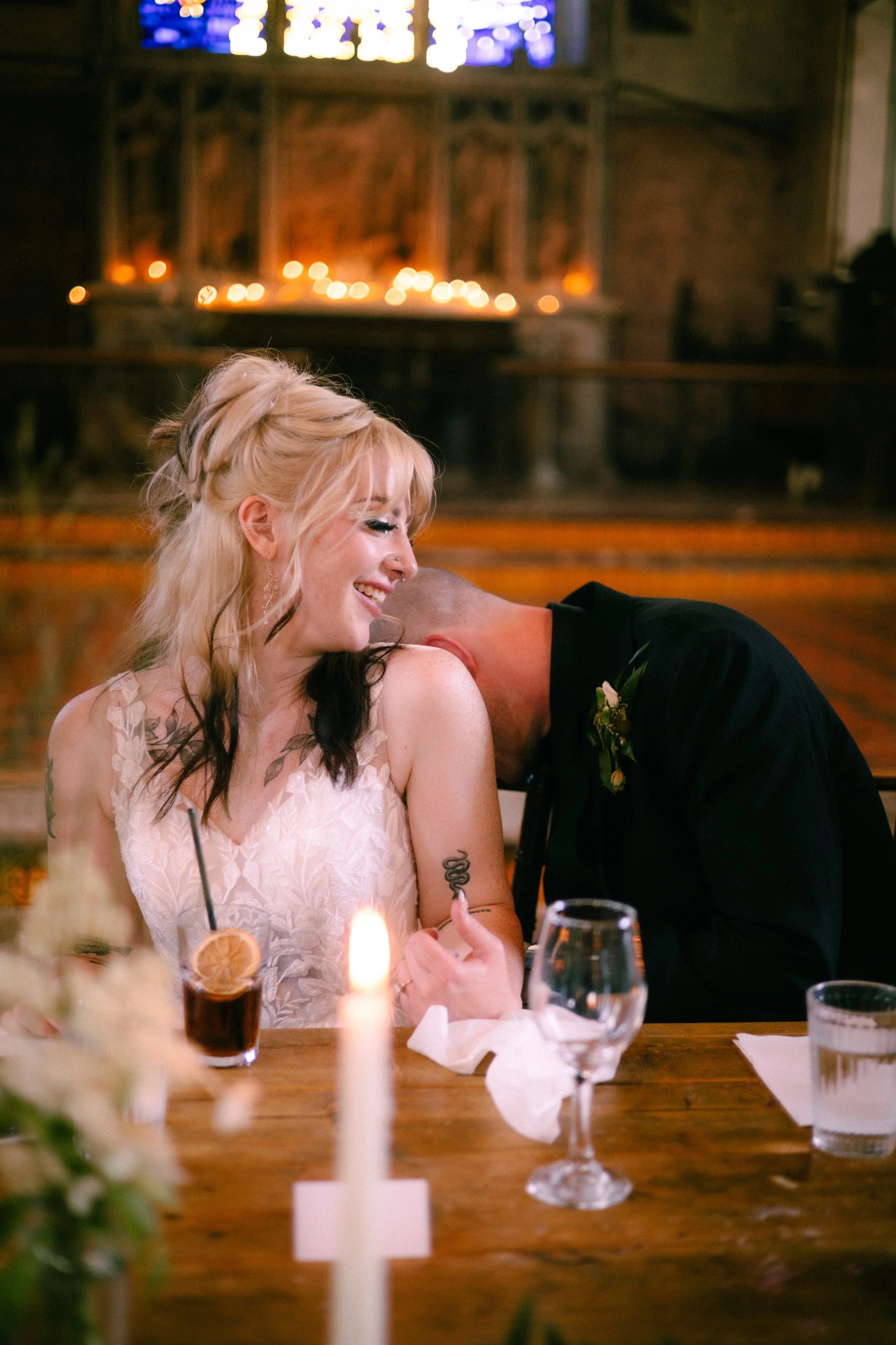 A woman with blonde hair smiling and a man with a shaved head leaning close, both seated at a table during a celebration in a dimly lit venue with candles and stained glass window in the background.
