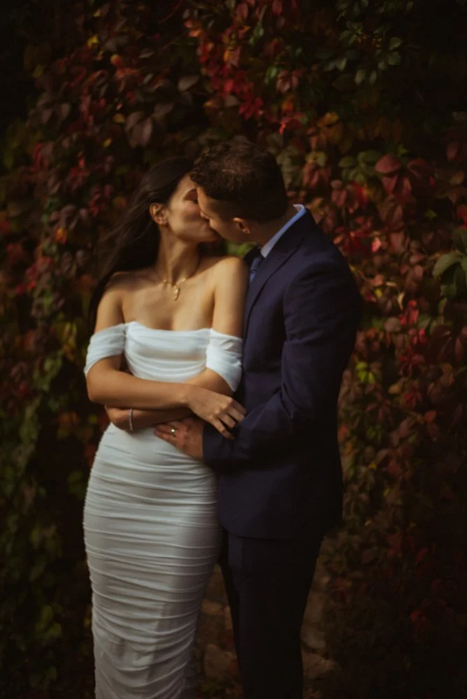 A couple sharing a kiss in front of a vine-covered wall, with the woman wearing a white off-the-shoulder dress and the man in a dark suit.