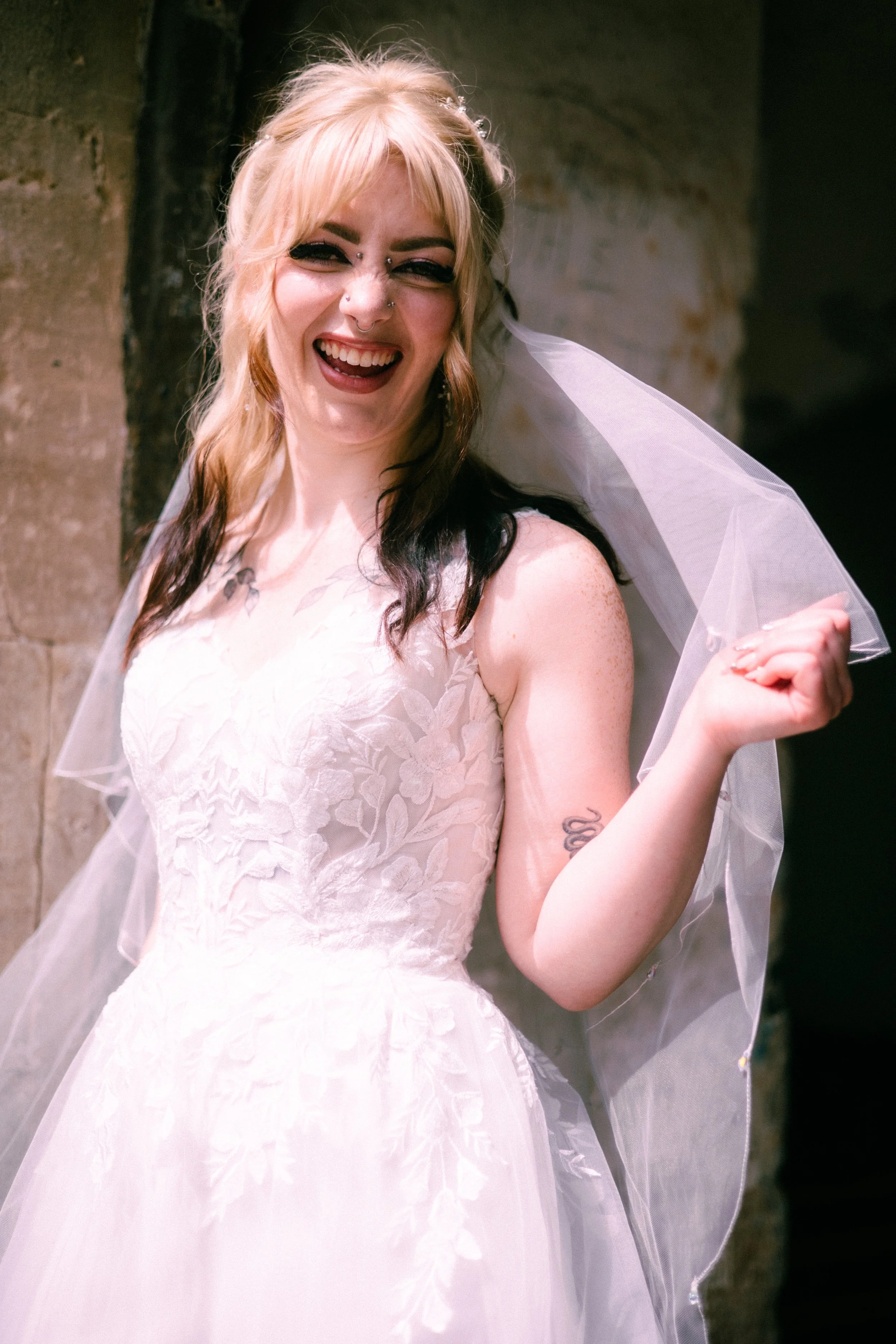 A woman with blonde and dark hair wearing a white lace wedding dress and a veil, smiling and holding up her arm with a tattoo, standing against a stone wall.
