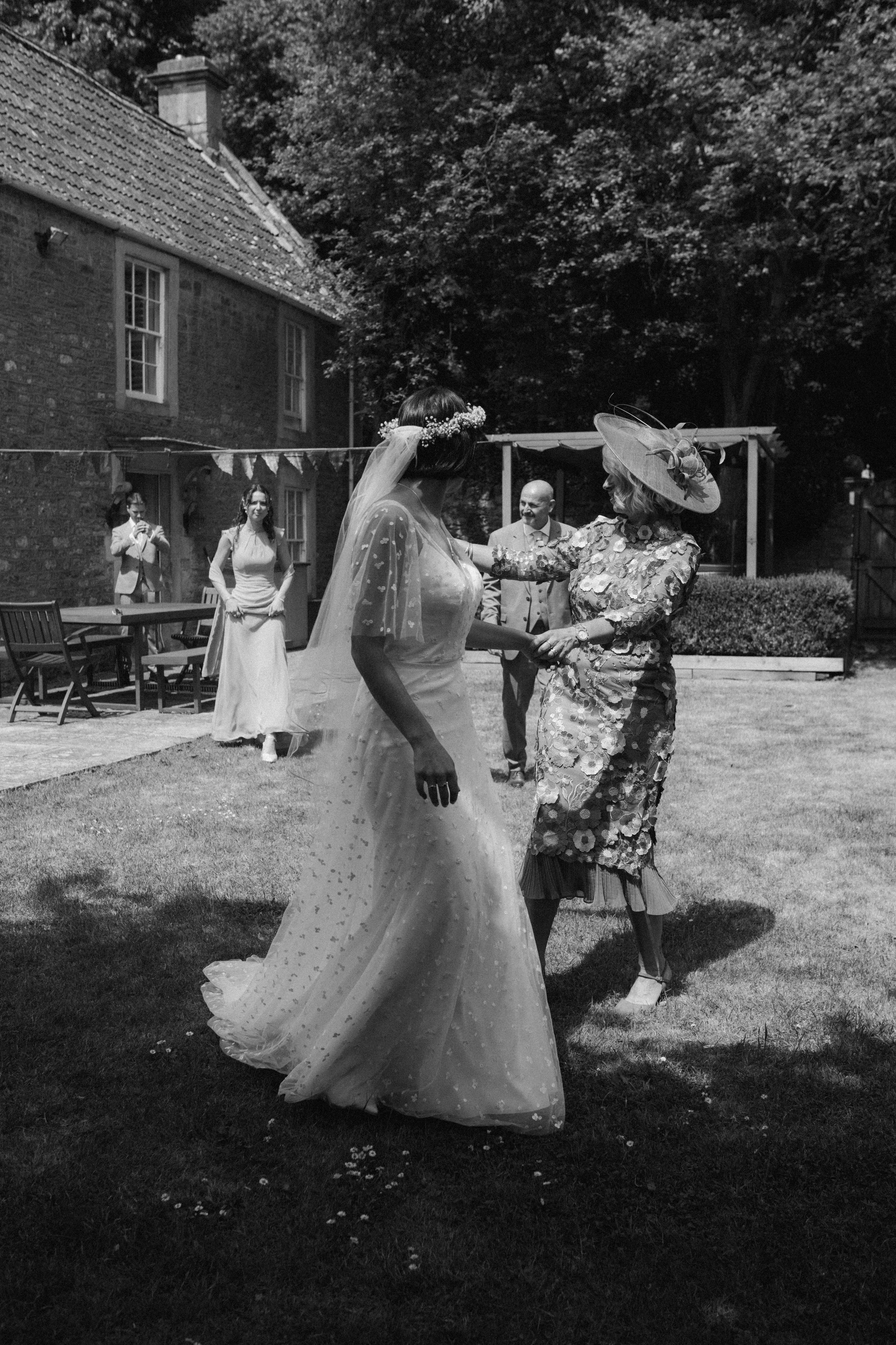 A bride in a wedding dress and veil dancing with an older woman wearing a large hat and floral dress during a wedding celebration outdoors, with guests and a brick building in the background.