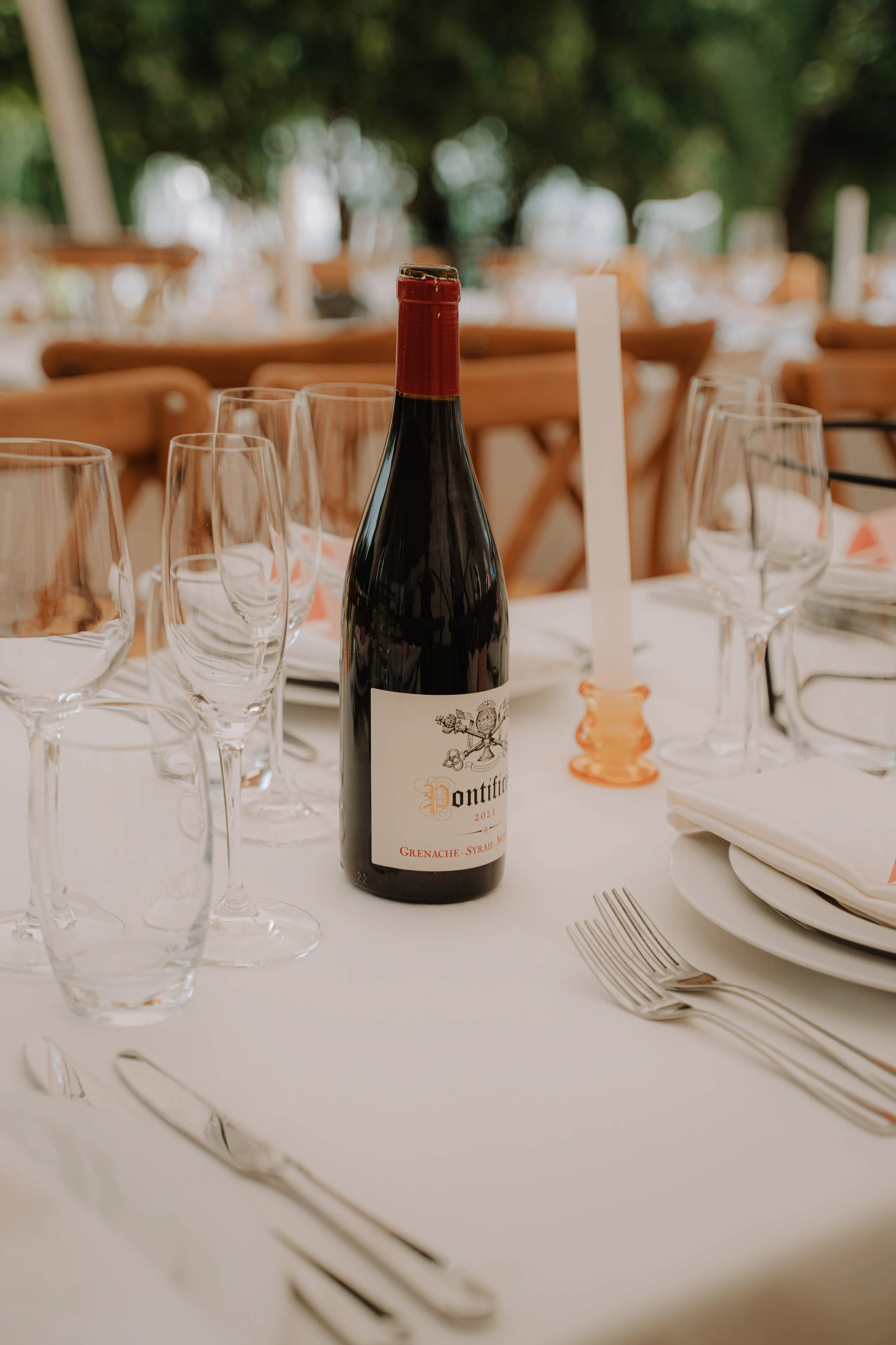 A wine bottle labeled 'Pontific' on a dining table set with several empty wine glasses, a white candle in an orange holder, lamborghini, a plate with a napkin, silver cutlery, and a white tablecloth, with a blurred outdoor background.
