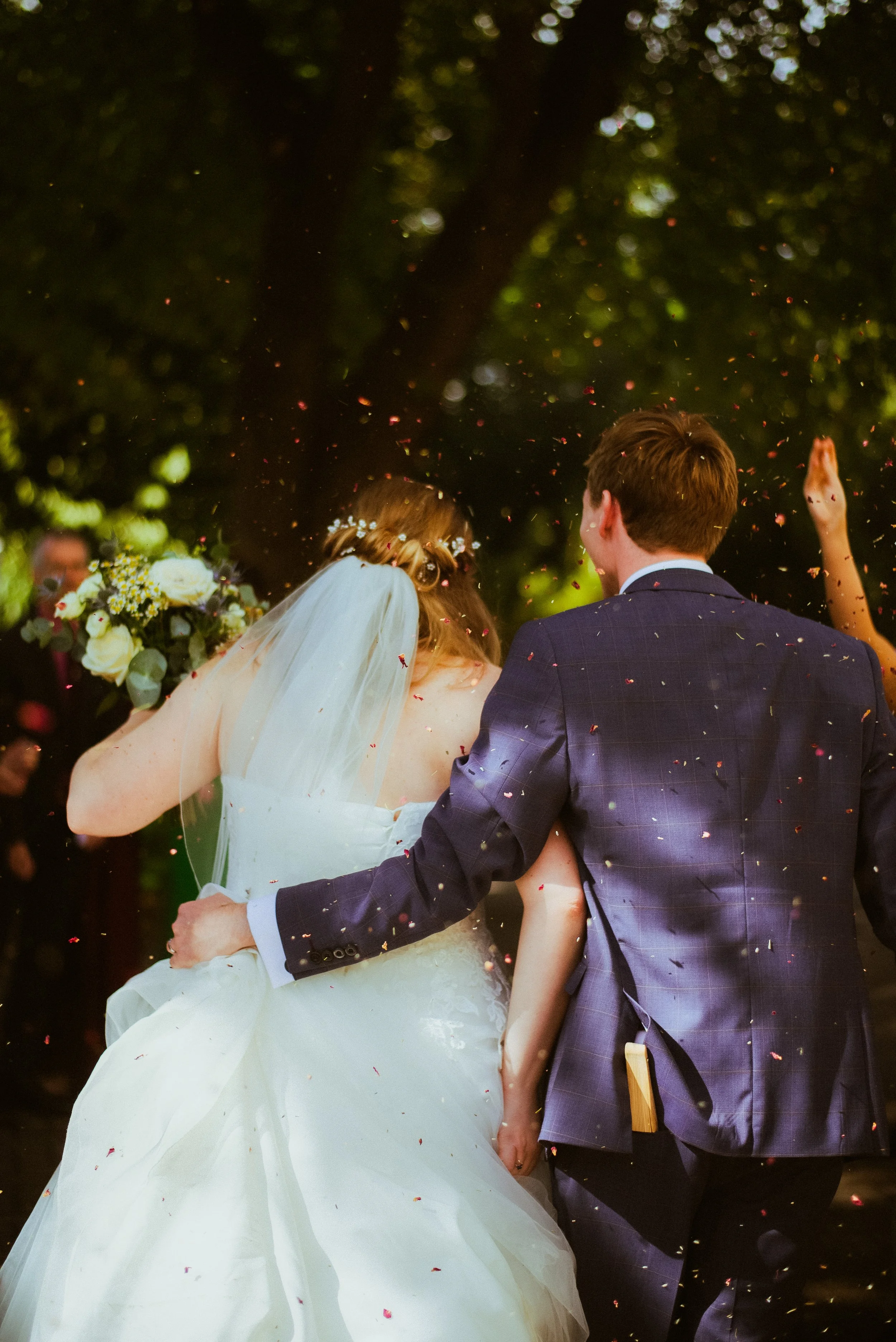 A bride and groom celebrate their wedding outdoors amidst falling confetti, with the bride holding a bouquet of flowers and the groom wearing a suit, during daytime.