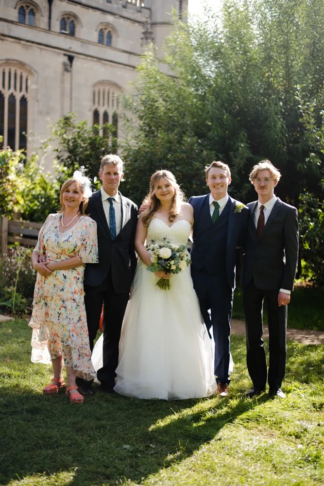 Group of five people, including a bride and groom, standing outdoors in front of a historic building and trees, dressed in wedding attire on a sunny day.