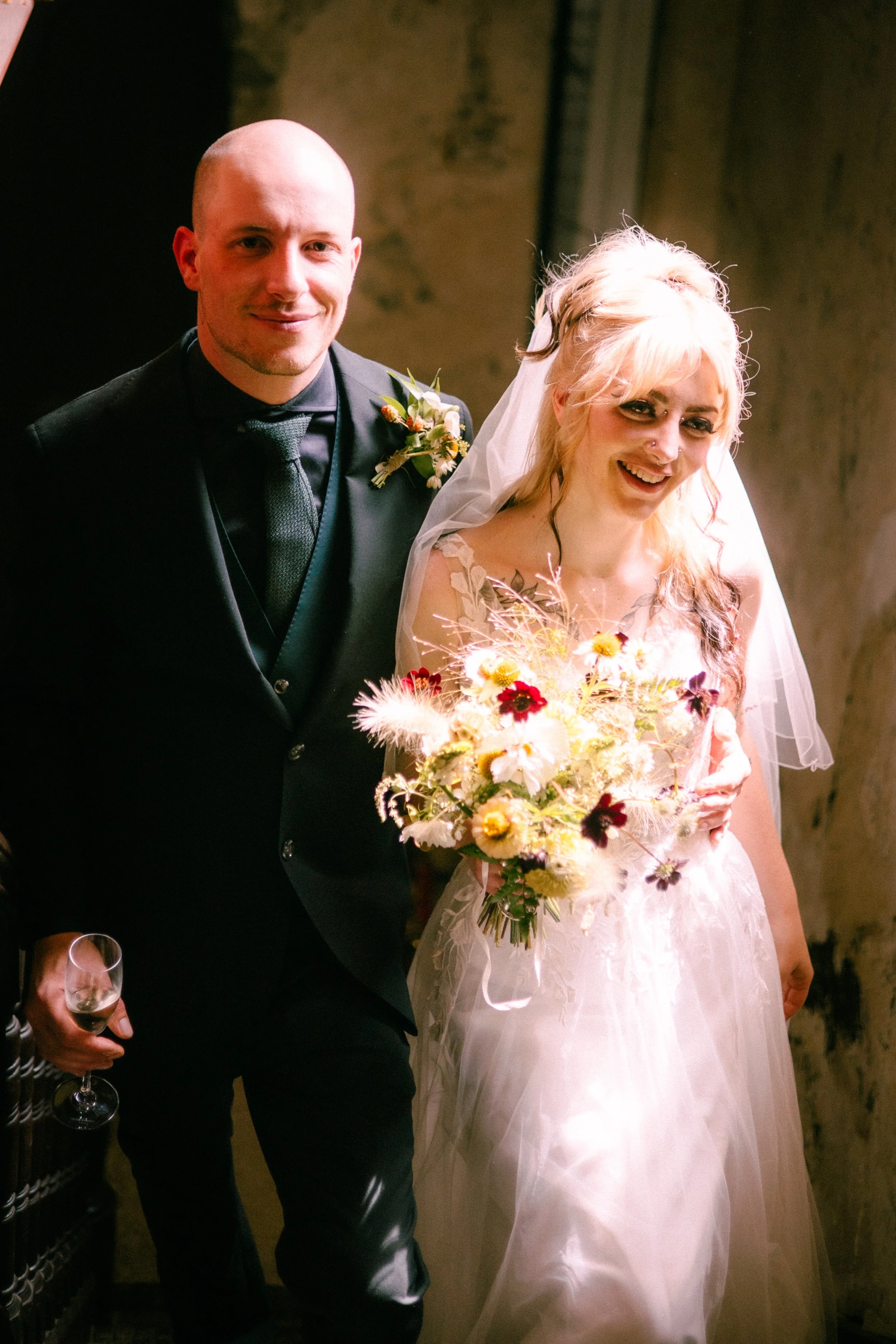 A bride and groom standing together at their wedding, with the bride holding a large bouquet of flowers and the groom holding a glass of wine.