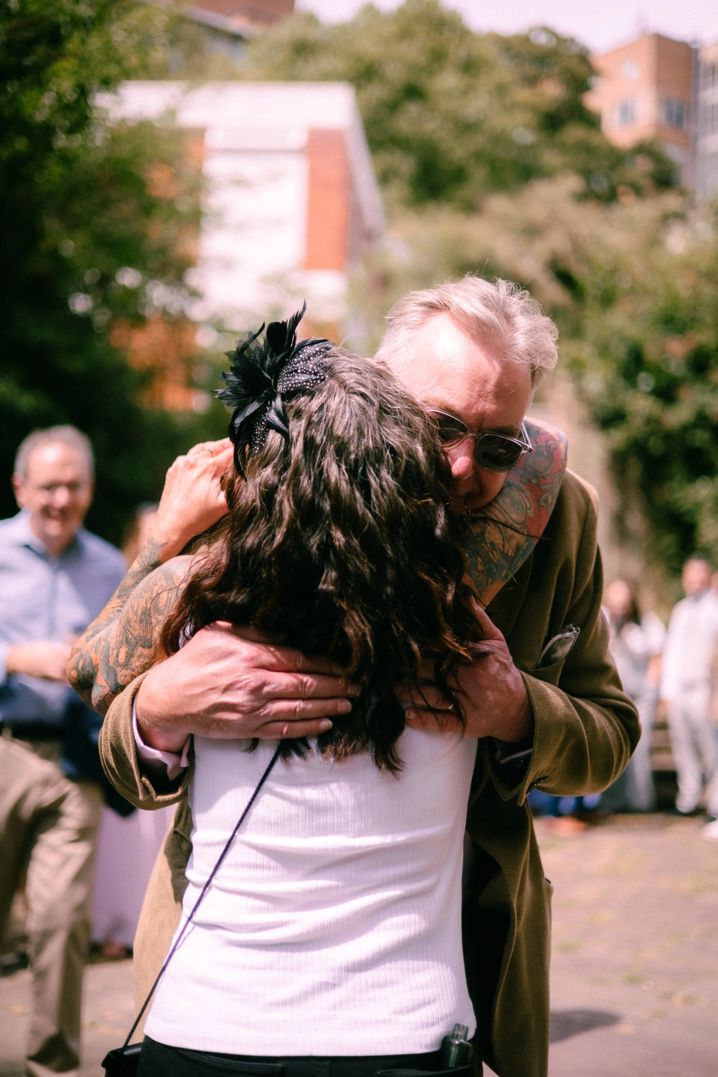 A man with gray hair and sunglasses hugging a woman with dark, curly hair and a black feathered hair accessory in an outdoor setting with trees and buildings in the background.