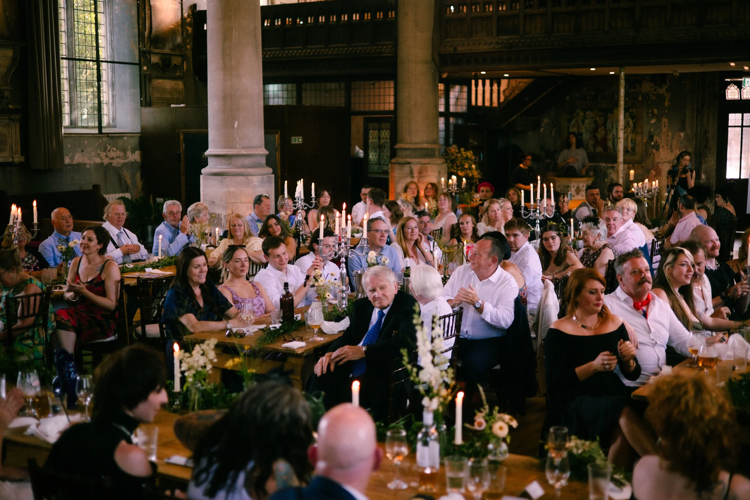 A large group of people sitting at decorated tables in a historic, dimly lit hall with high ceilings and large windows, attending a formal event or celebration.