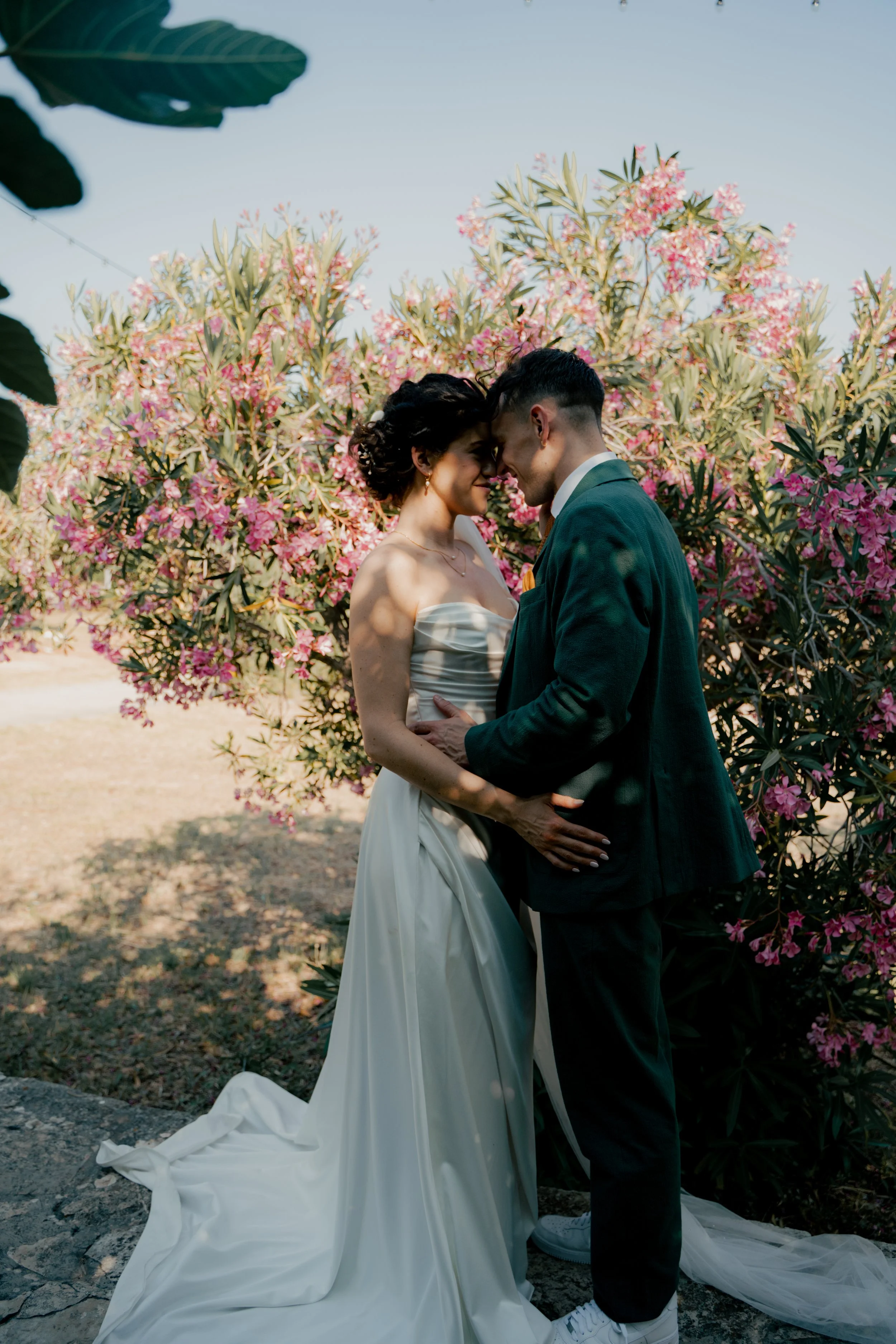 A bride and groom standing close together, touching foreheads and holding hands, outdoors with pink flowering bushes in the background.