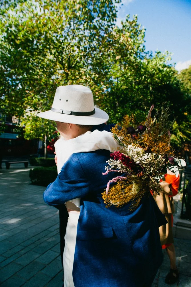 Person wearing a white hat and a blue suit hugging a child with a bouquet of flowers on their back outside on a sunny day.