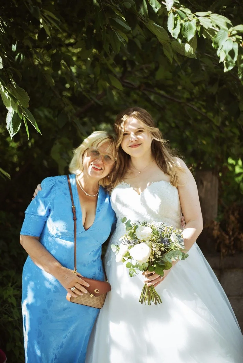 A young woman in a white wedding dress holding a bouquet standing next to an older woman in a blue dress under leafy trees.