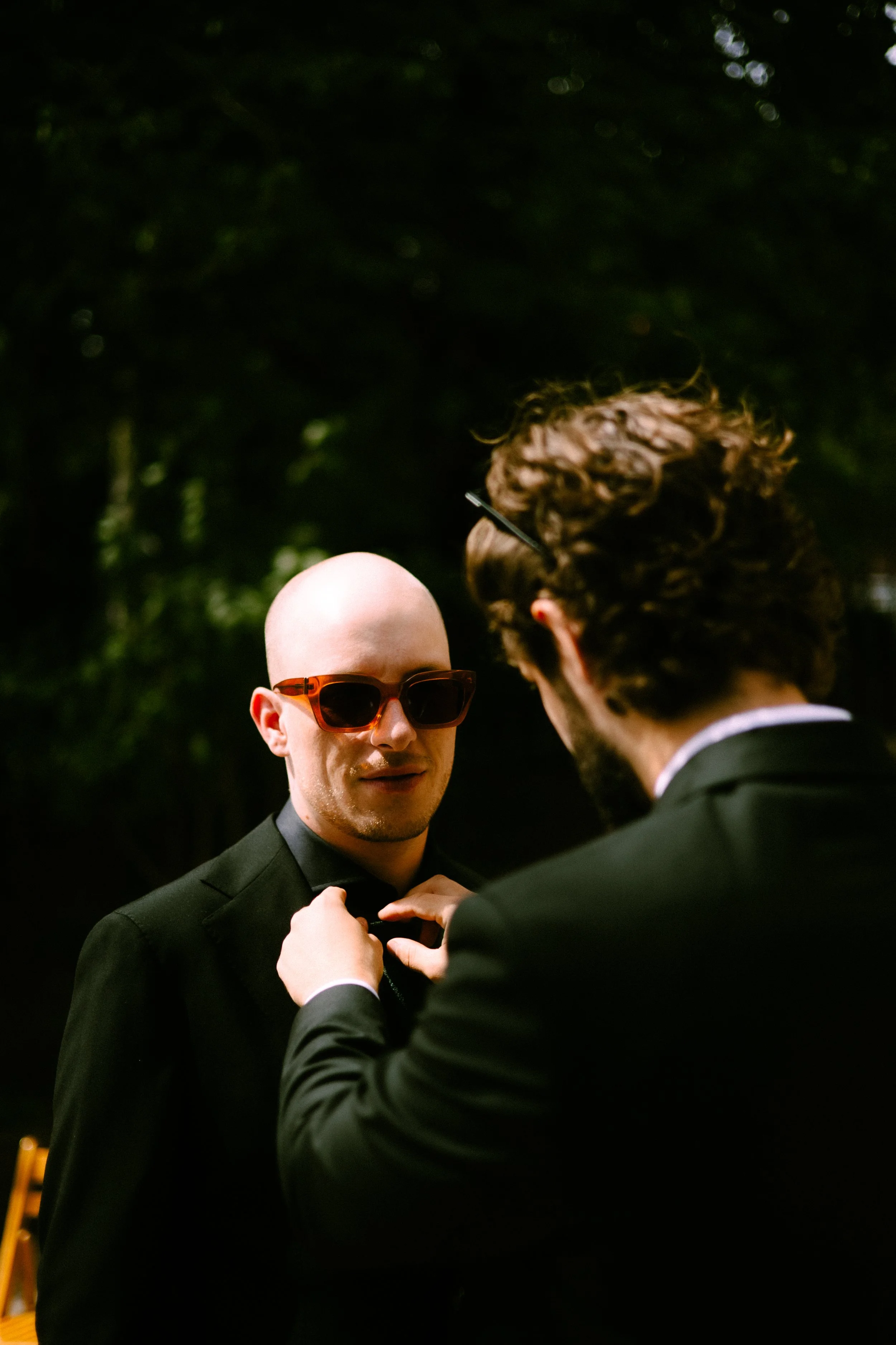 Two men in tuxedos, one adjusting the other's bow tie, outdoors with a dark green leafy background.