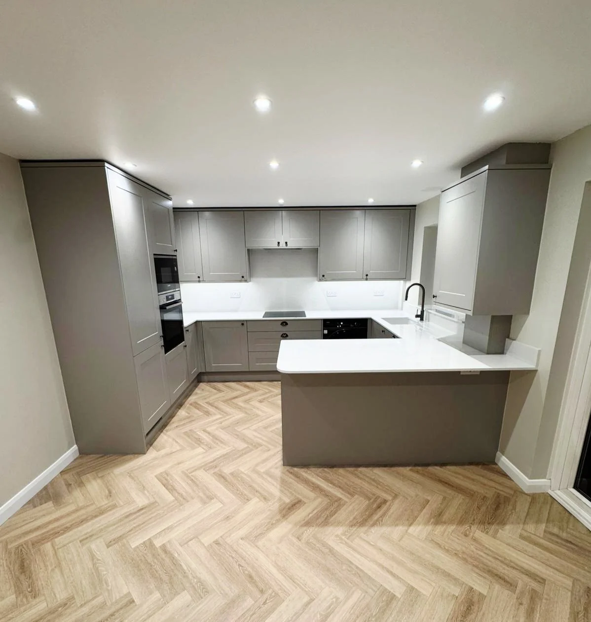 Warm taupe grey shaker kitchen installation in Camberley with white quartz worktops, full-height quartz splashbacks, island-style peninsula and integrated appliances.