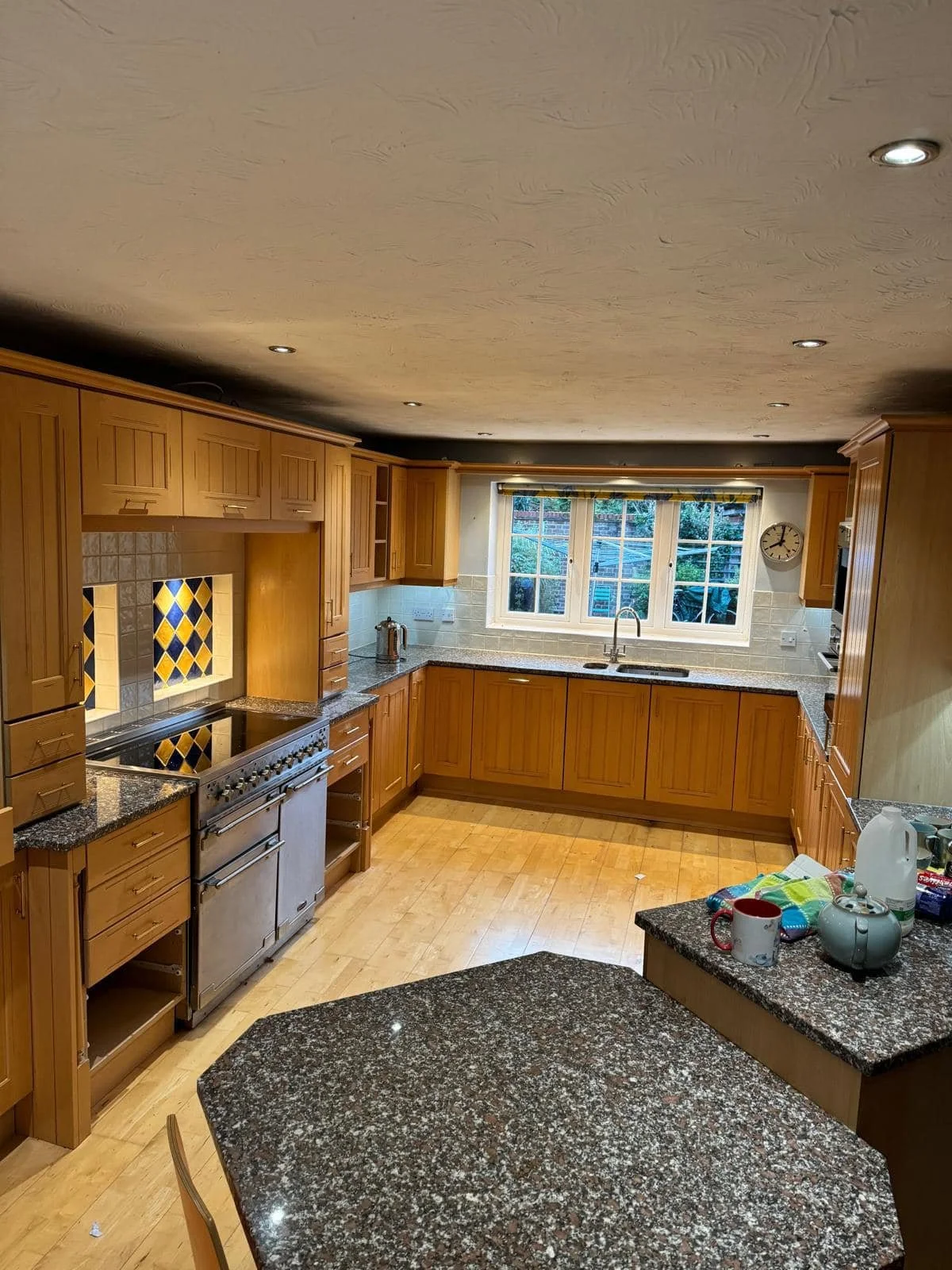 A spacious L-shaped kitchen with wooden cabinets, granite countertops, a stove with a decorative tile backsplash, and a large window above the sink. The kitchen has wood flooring and recessed lighting in the ceiling.