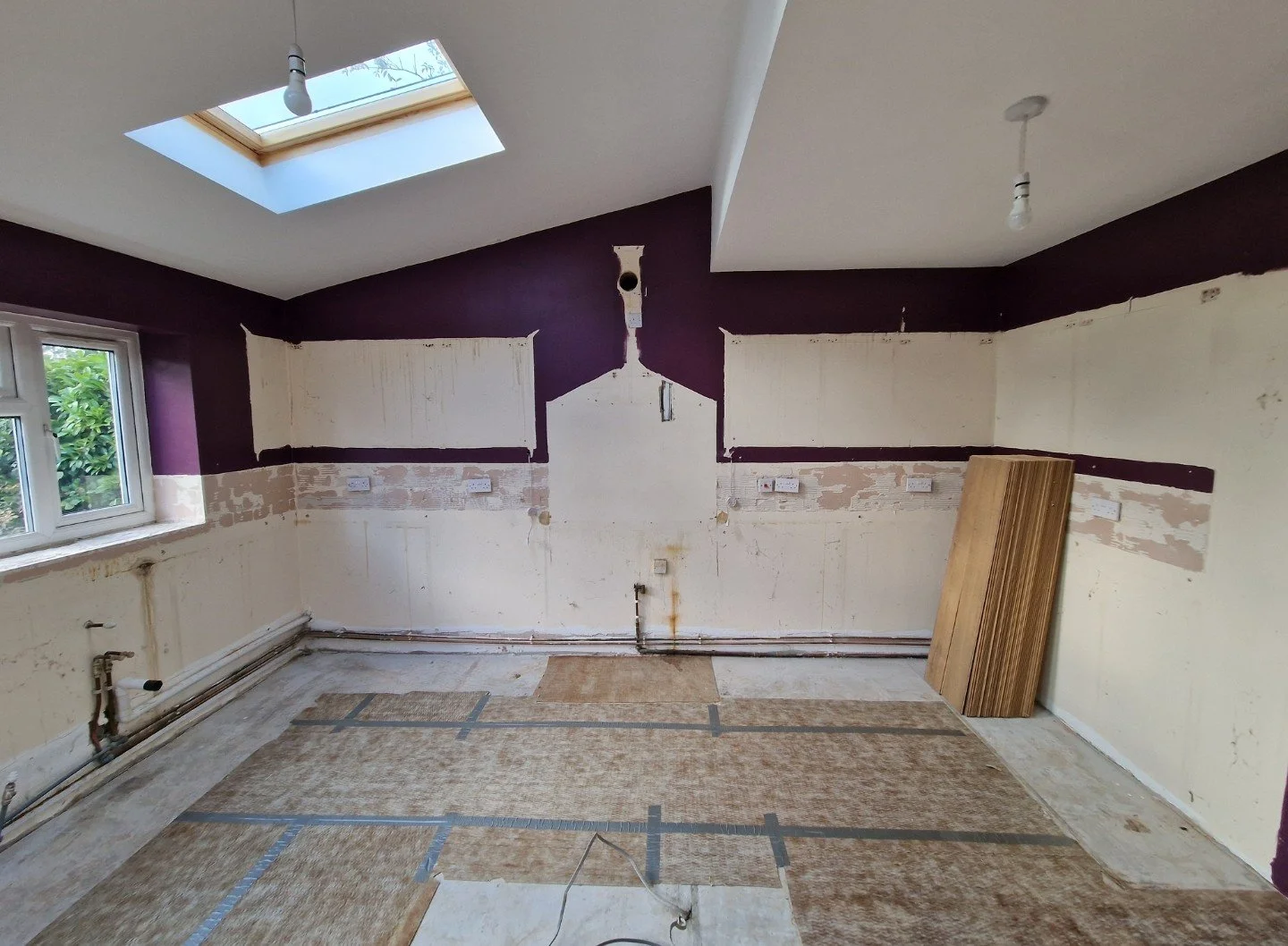 Kitchen renovation before installation with partially painted walls, a window, and flooring covered with protective sheets, a stack of wooden planks leaning against the wall, and ceiling lights.