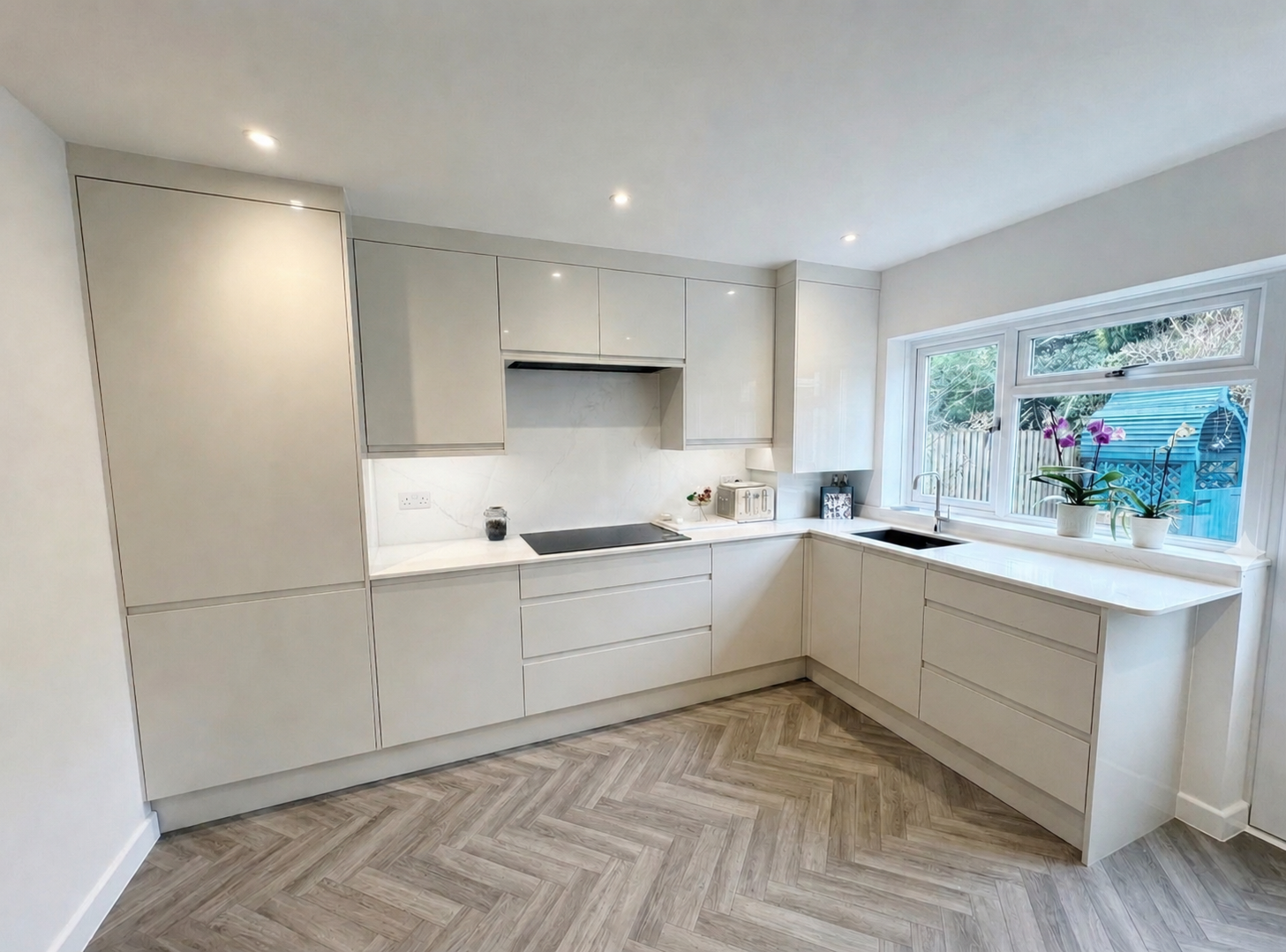 Gloss handleless kitchen in Sunningdale with marble-effect worktops, integrated hob, sink under the window, tall storage units and herringbone LVT flooring by Blacktail Kitchens.