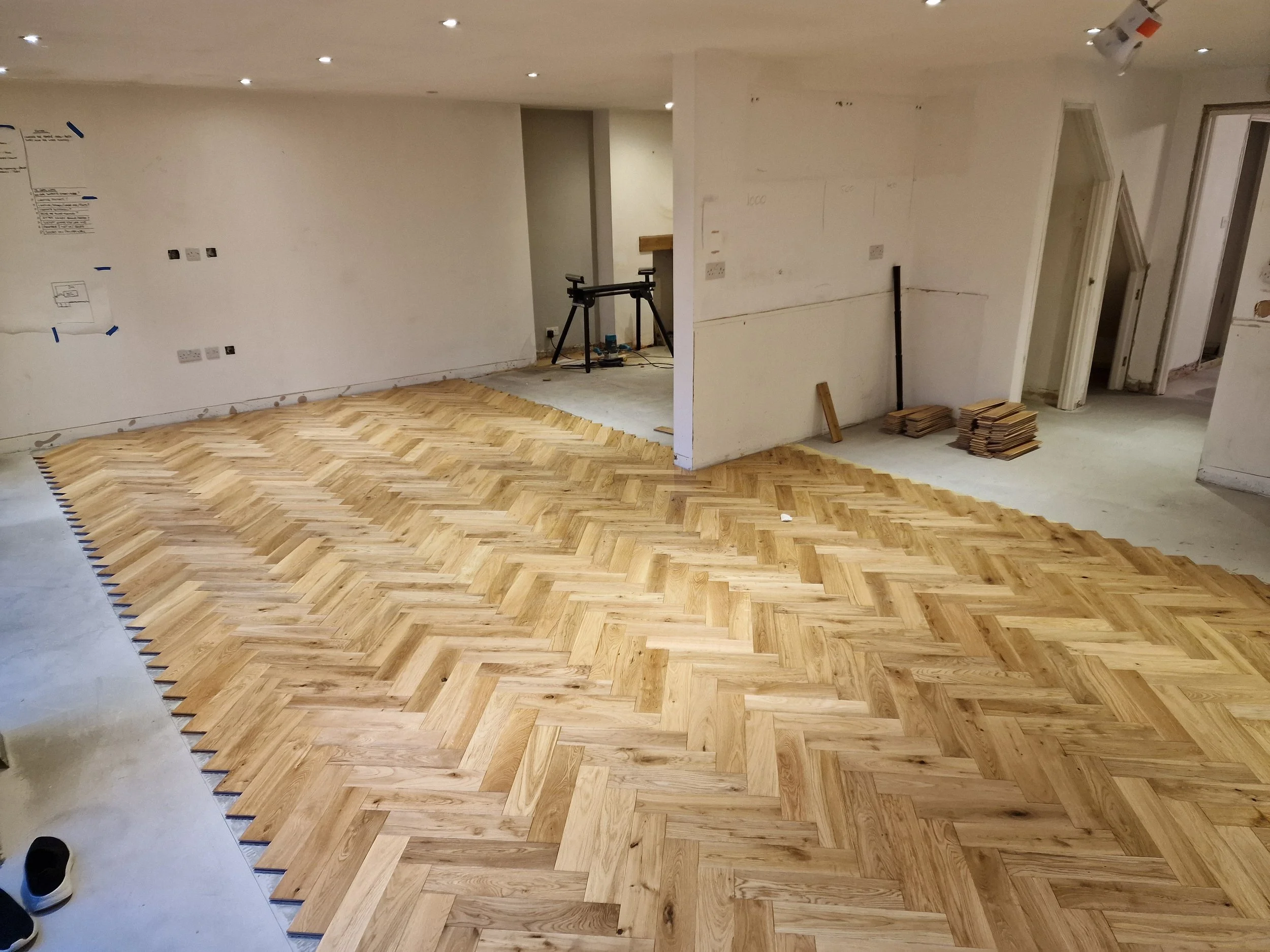 Kitchen renovation before installation with a newly installed herringbone pattern hardwood floor, and construction tools and materials scattered around.