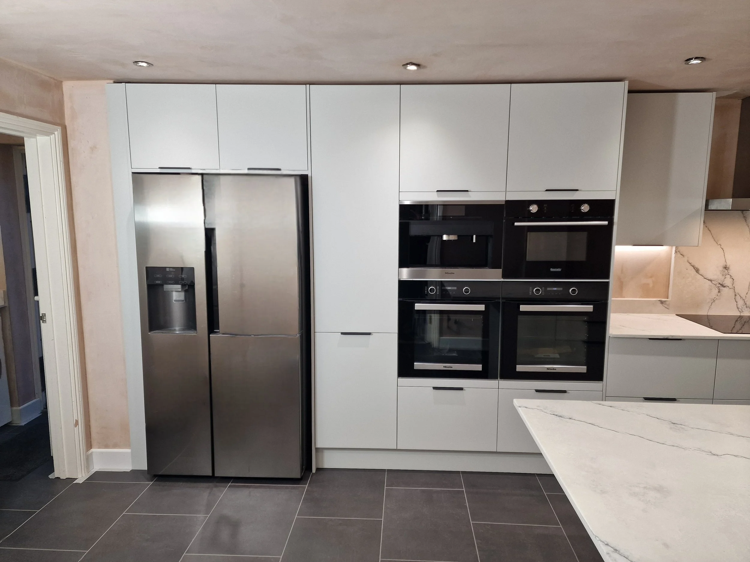 Modern kitchen with stainless steel refrigerator, built-in ovens, microwave, and sleek gray cabinetry, marble countertops, and dark tiled flooring.
