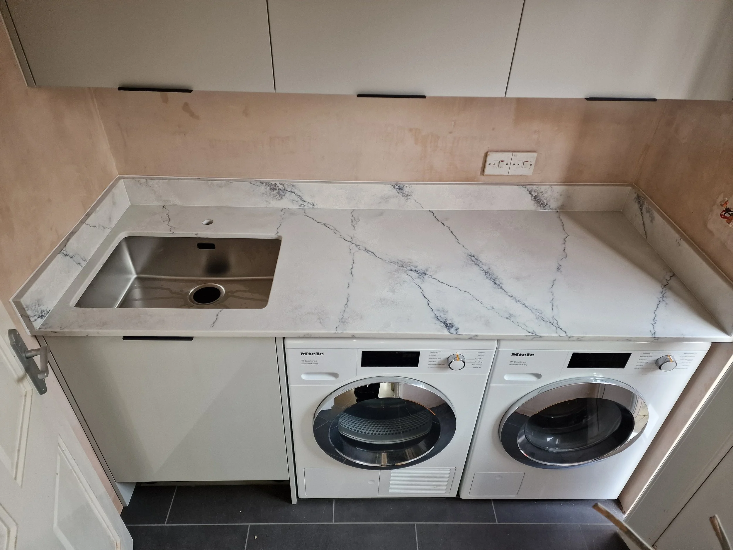 A laundry room with a white marble countertop, a built-in stainless steel sink, a washing machine, and a dryer underneath the counter. The walls are painted a light beige color, and there's an electrical outlet and a power switch on the wall.