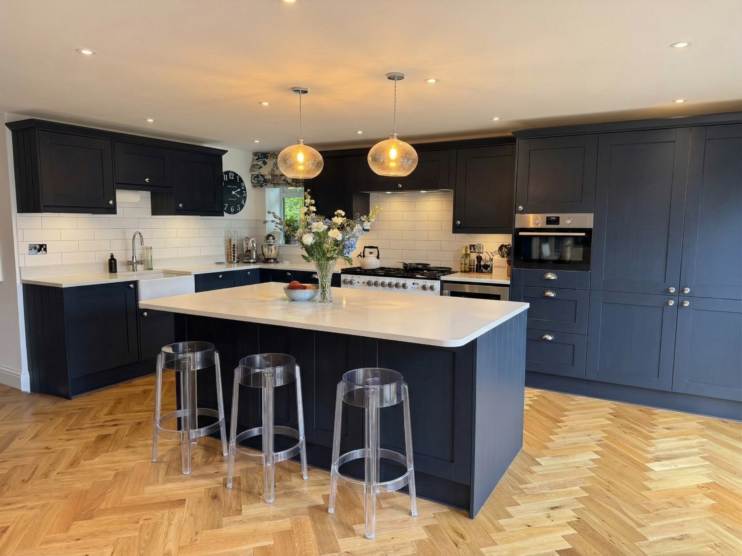 A modern kitchen with dark blue cabinets, white subway tile backsplash, and a white island with a vase of flowers on top. There are pendant lights hanging from the ceiling and clear barstools at the island. Hardwood flooring covers the floor.