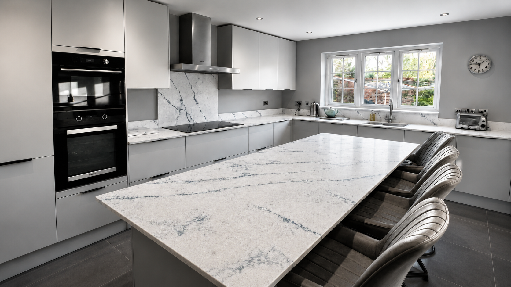 Modern grey kitchen installation in Sunningdale with a large quartz island, integrated ovens, induction hob, marble-effect splashback and grey tiled flooring by Blacktail Kitchens.