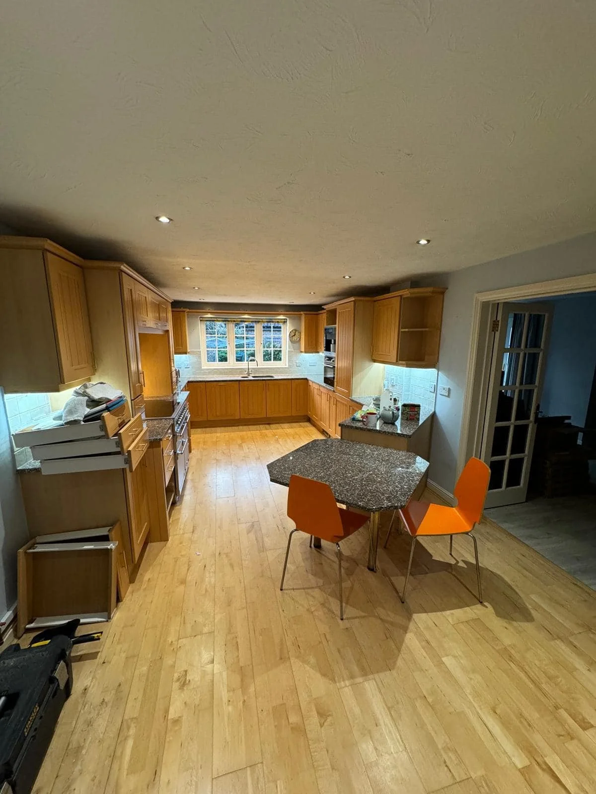 Dated wooden cabinets, a granite island, and a window above the sink. Two orange chairs are placed around a small, variably shaped countertop. The floor is made of light wood, and the ceiling has multiple recessed lights.