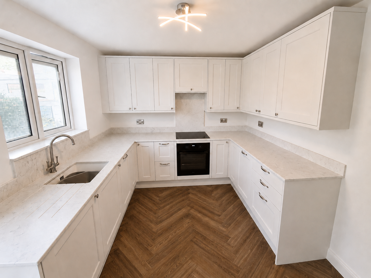White shaker kitchen in Ascot with marble-effect worktops, integrated appliances, sink area and warm herringbone flooring by Blacktail Kitchens.