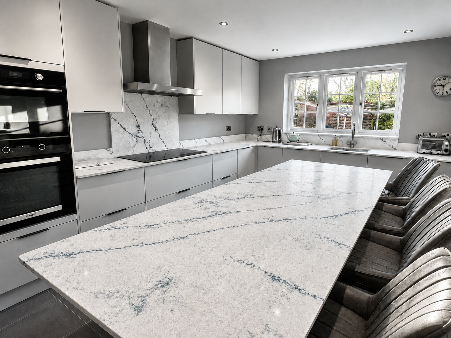 Modern kitchen with white quartz and blue vein countertops, gray cabinets, built-in oven and stove, stainless steel range hood, large window, clock, and bar stools.