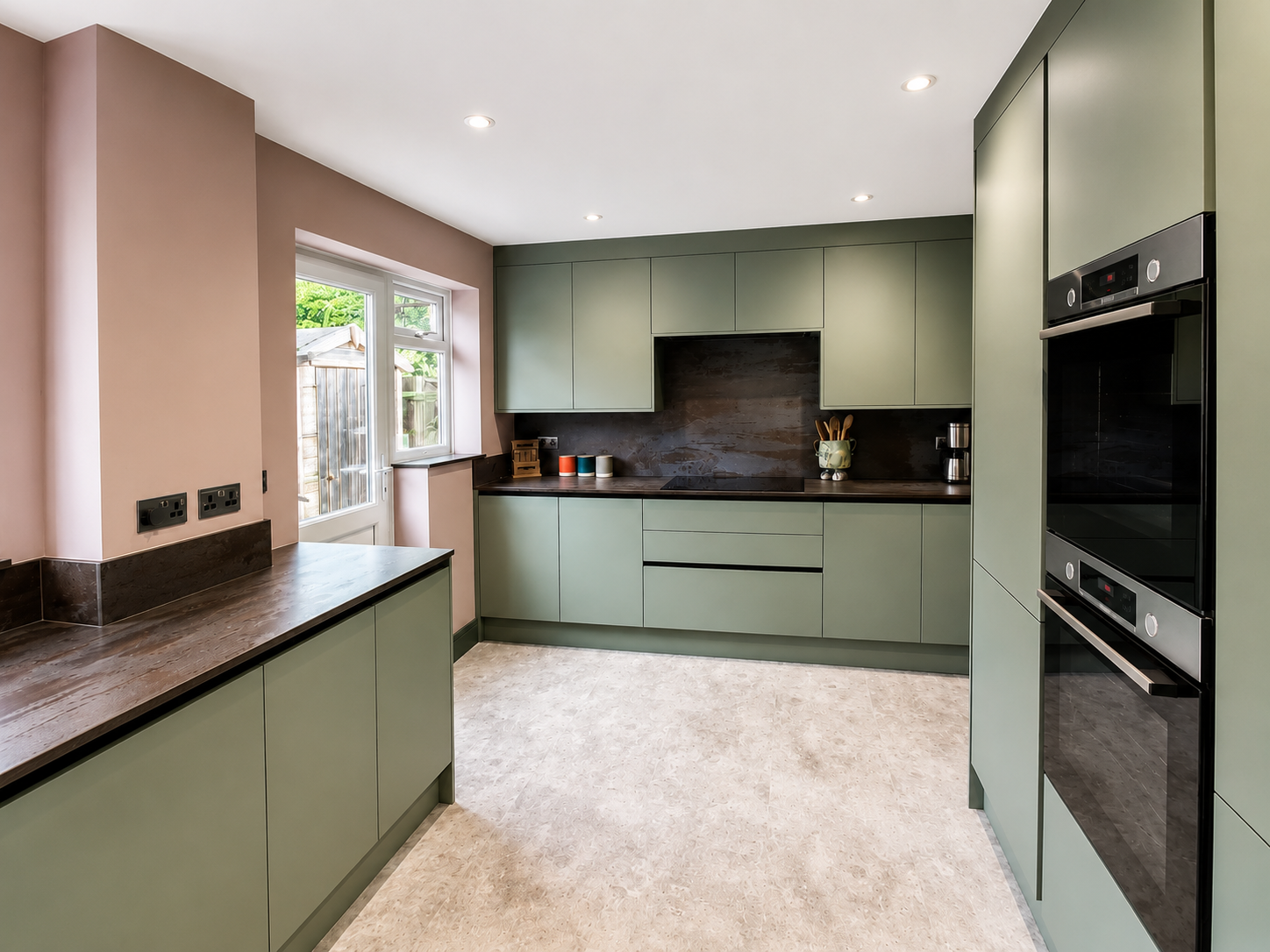 Modern Warfield kitchen with true handleless green units, dark quartz worktops, matching splashback, integrated hob area and warm pink walls.