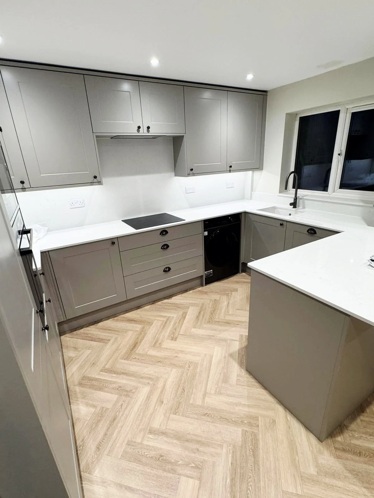 Completed taupe grey fitted kitchen in Camberley with shaker units, white quartz worktops, black sink and tap, microwave housing and herringbone LVT flooring.