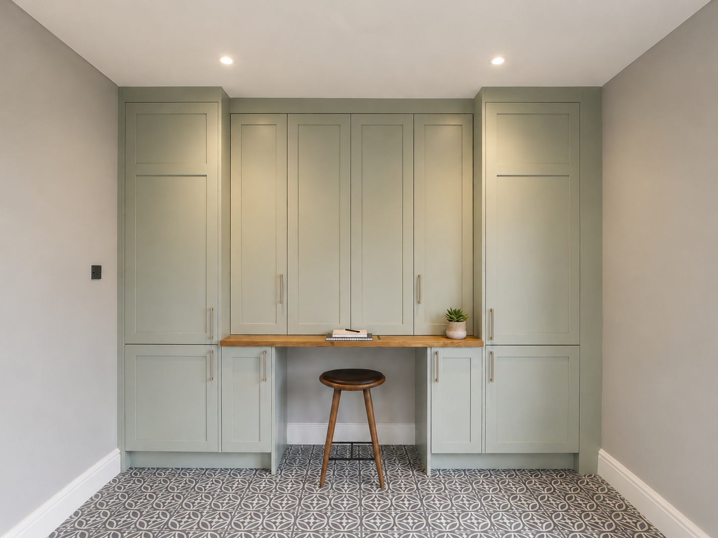 Bespoke fitted workspace in Camberley with sage green storage units, oak desktop, bi-folding wall unit doors and patterned floor tiles.