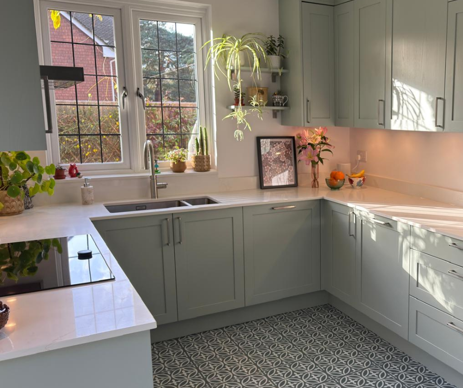 A bright kitchen with light green cabinets, a white marble countertop, and a window with a garden view. Decor includes potted plants, flowers, and framed art.