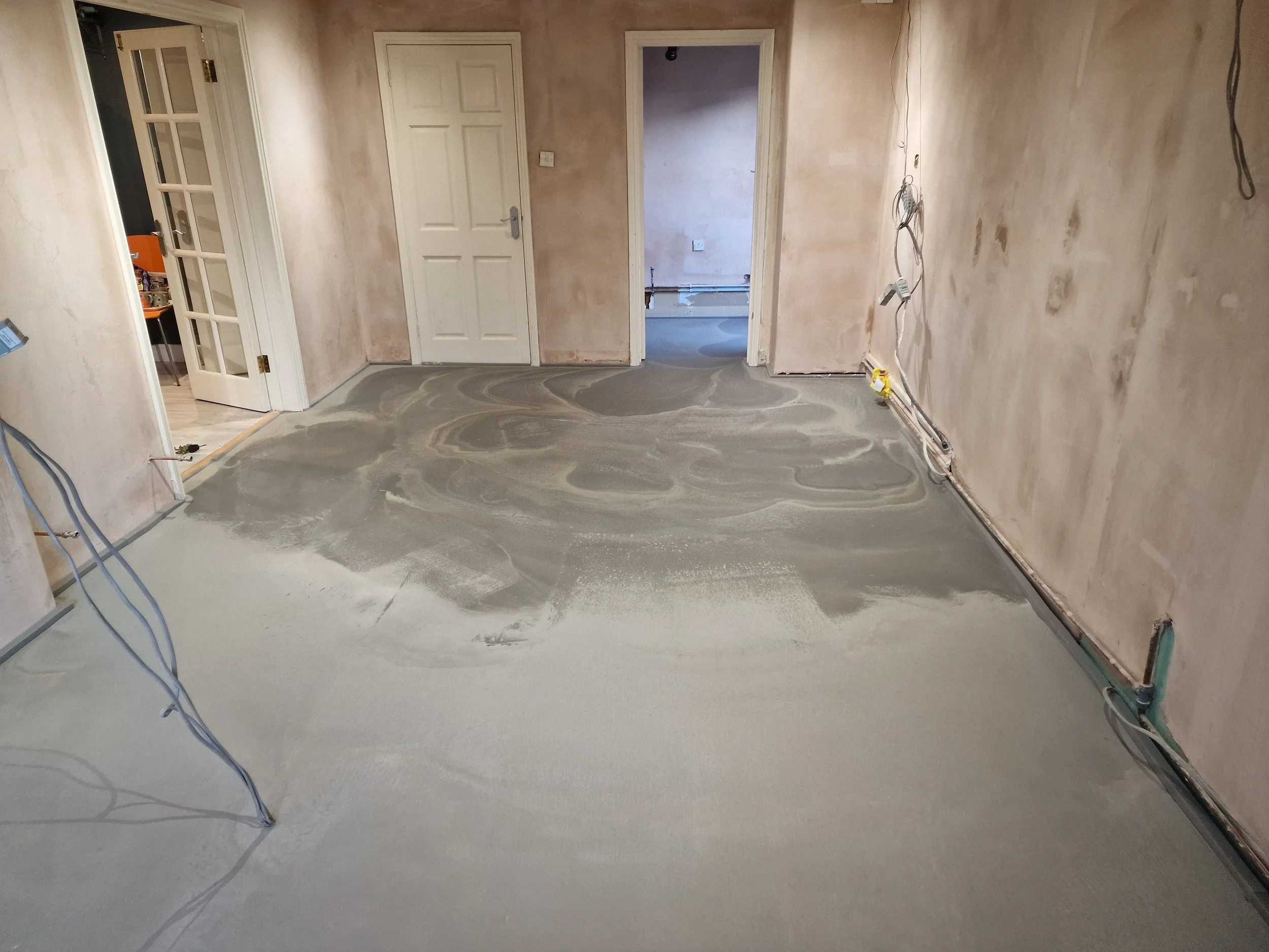 Kitchen preparation before installation with unfinished concrete floor, exposed wiring, and unpainted walls. A doorway leads to another room, and there are white doors and trim.