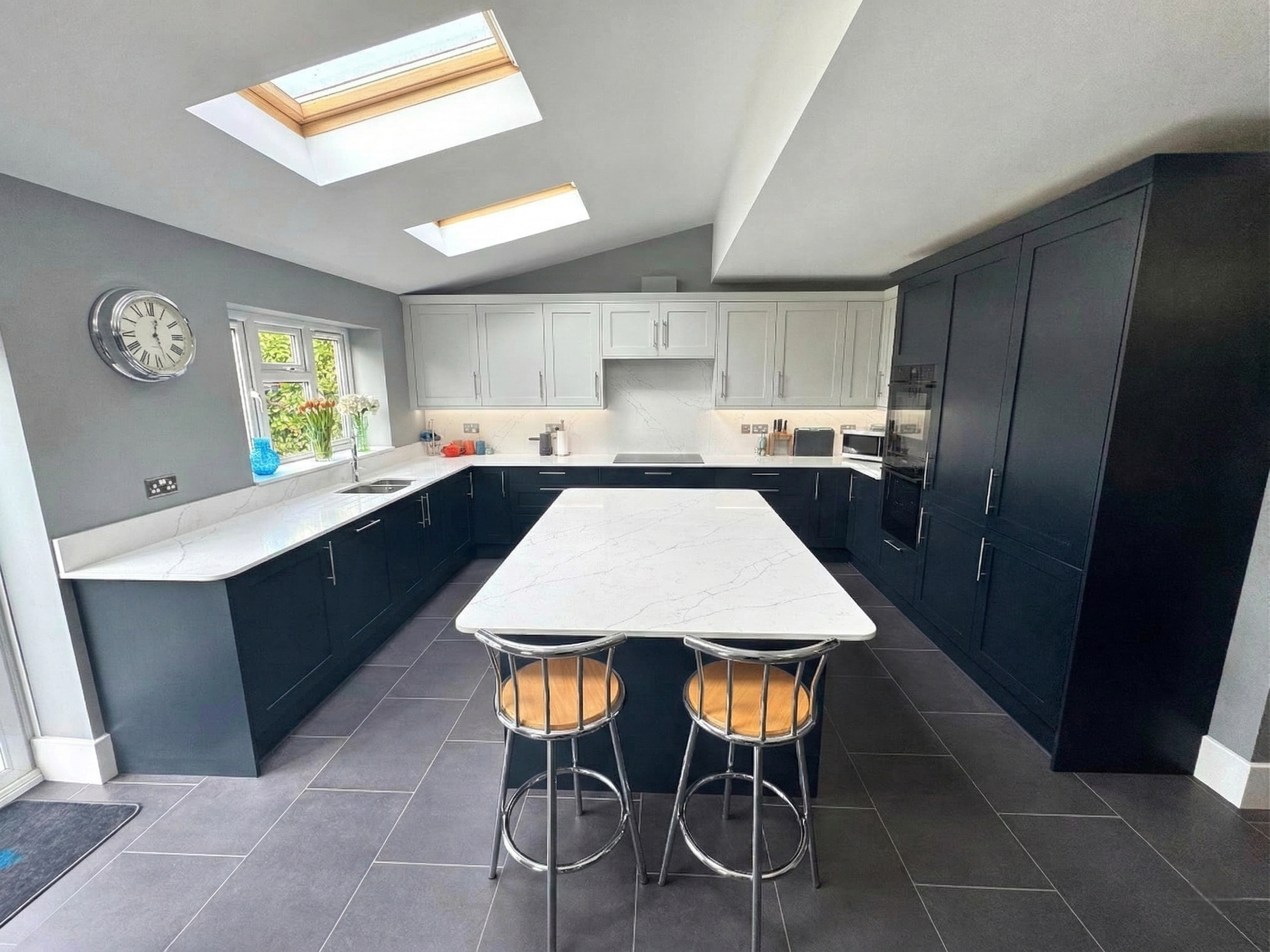 Modern kitchen with black lower cabinets, white upper cabinets, a white countertop, and a kitchen island with two metal barstools. Skylights in the ceiling provide natural light, and a wall clock is visible near a window.