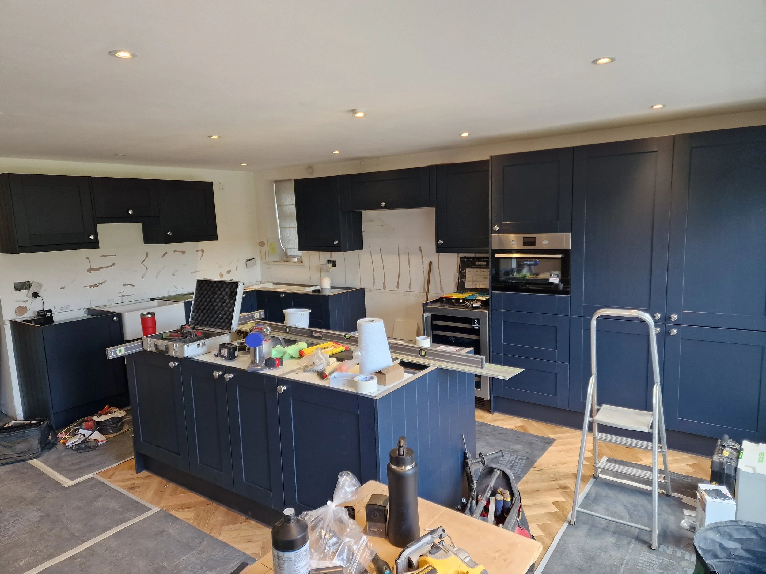 Kitchen renovation progress with dark blue cabinets, tools, and partially installed backsplash, and construction materials and ladder present.