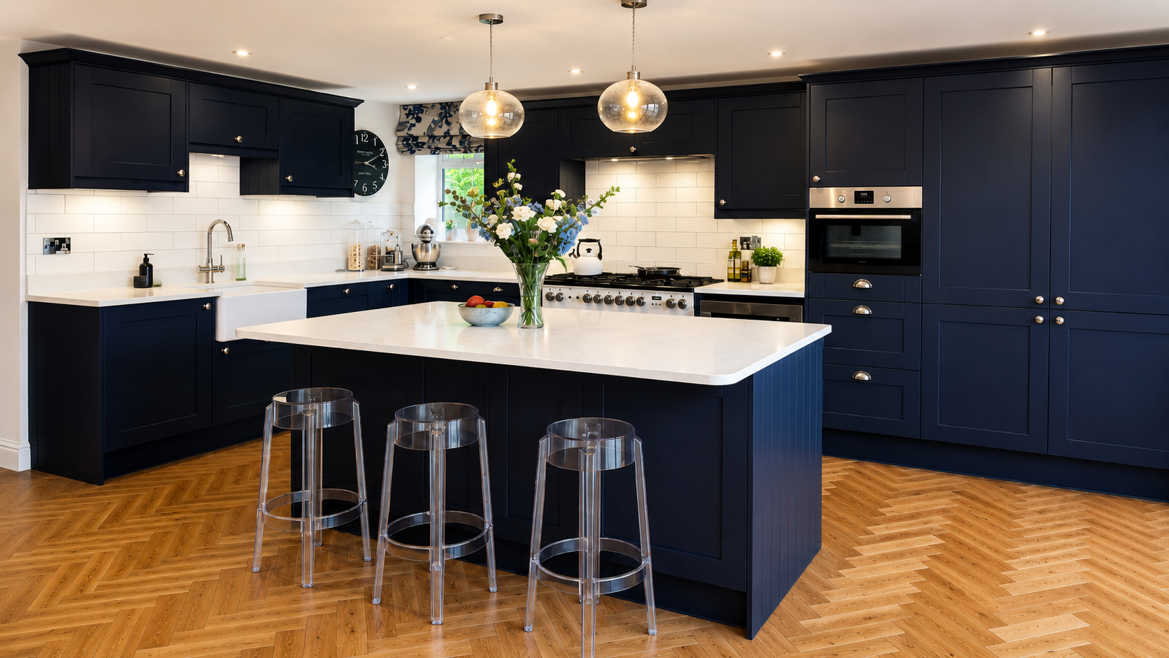 Navy shaker kitchen with a large white worktop island, pendant lighting, clear bar stools, range cooker and herringbone wood flooring by Blacktail Kitchens.