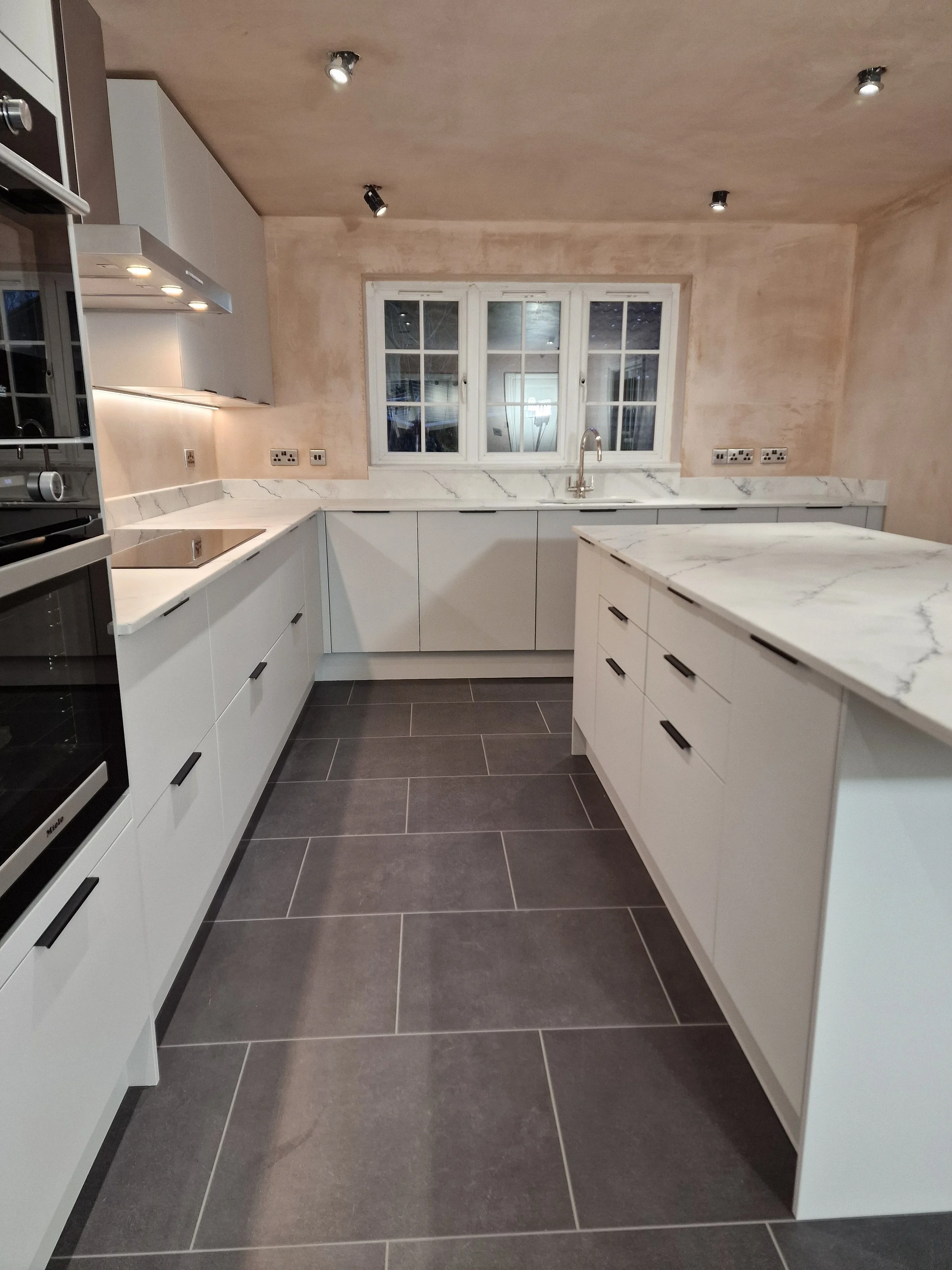 Modern kitchen with white cabinetry, marble countertops, large window above the sink, and dark gray tiled flooring.