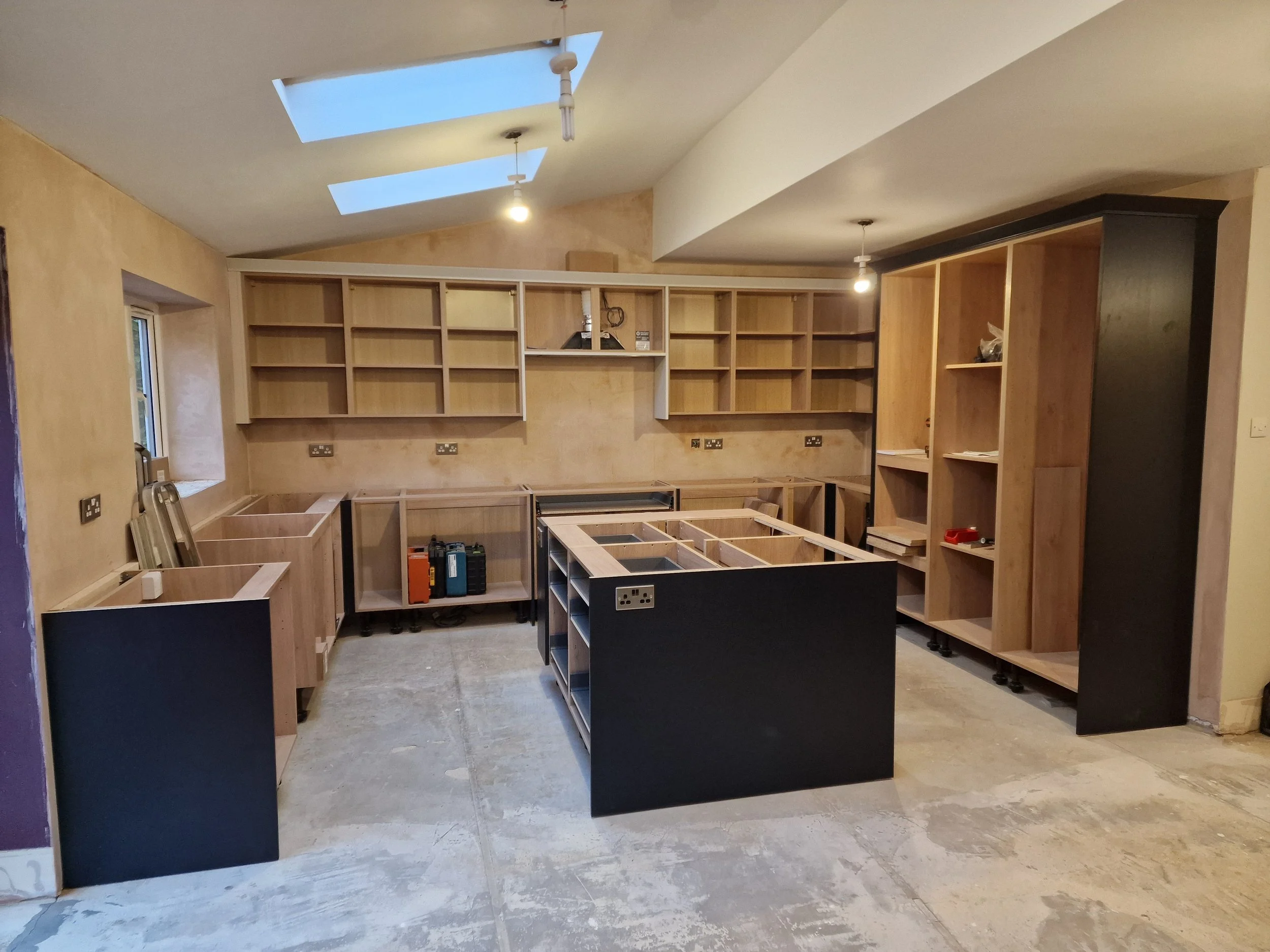 Room with unfinished wooden kitchen cabinets and shelving units, countertops not yet installed, ceiling with skylights, and construction tools on the floor.