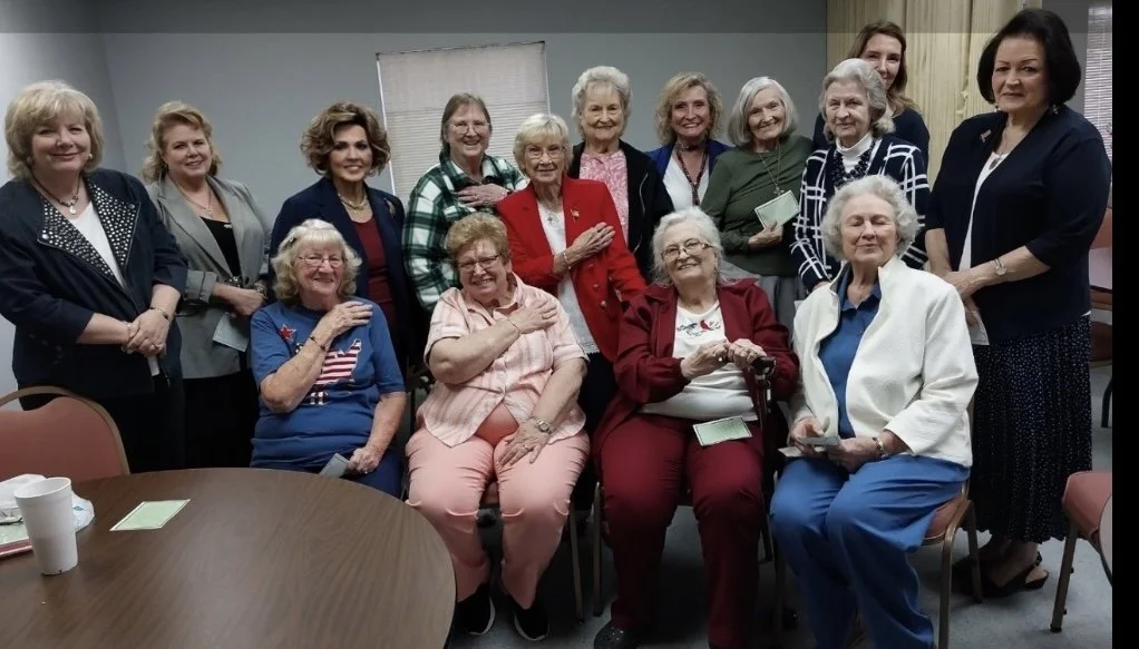 Group of elderly women and a few younger women gathered indoors, some seated and some standing, smiling for a photo.