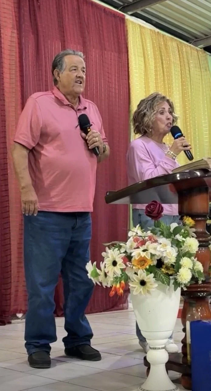 An elderly man and woman standing on a stage, speaking into microphones. The man is wearing a pink polo shirt and jeans, and the woman is wearing a pink top and blue jeans. There is a large flower arrangement on a white vase in front of them, with a rose and white flowers. The background has red and yellow curtains.