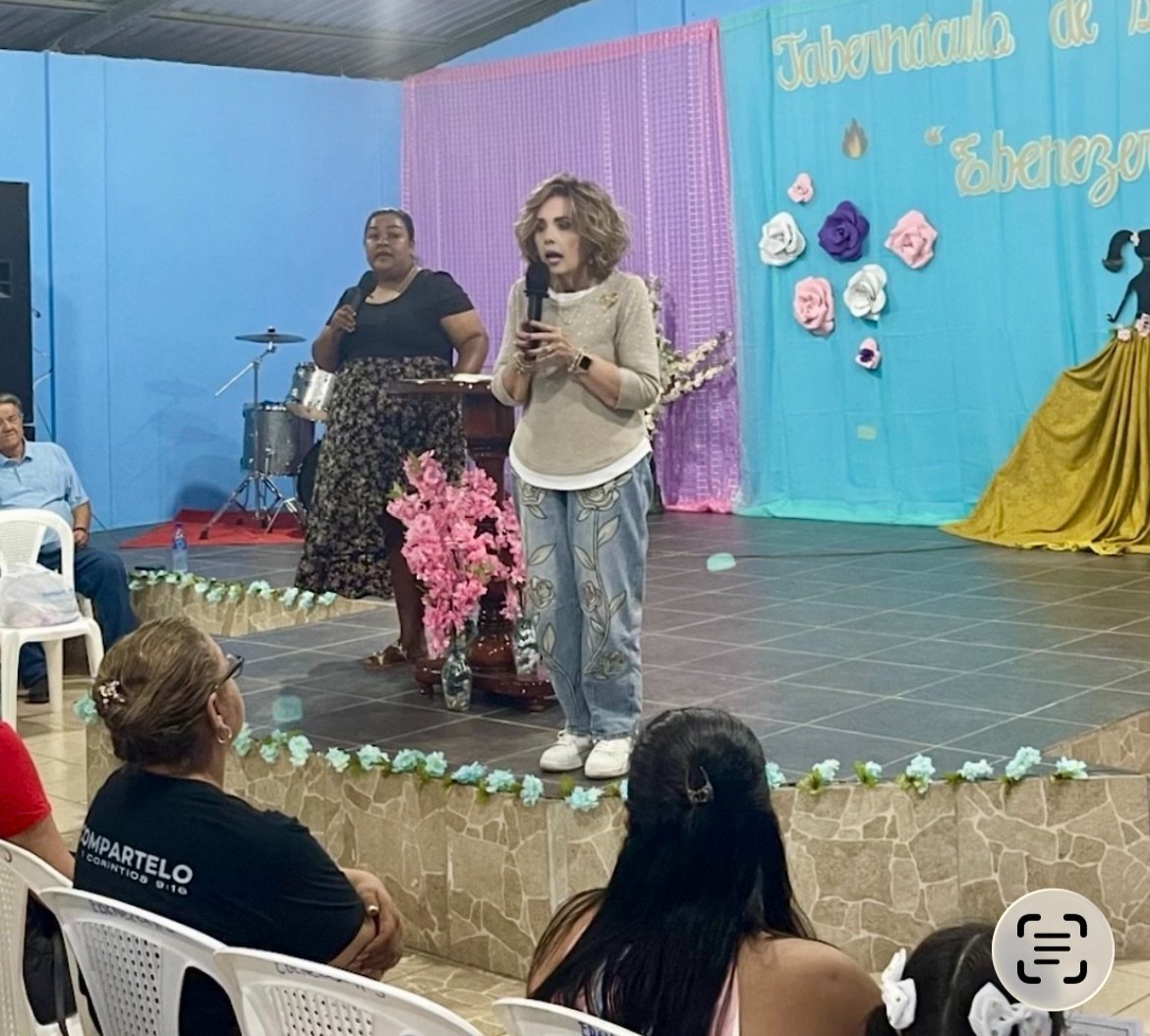 Women speaking on a stage decorated with pink and purple flowers, in front of an audience, with a blue backdrop and pink curtain, during a community event or church gathering.