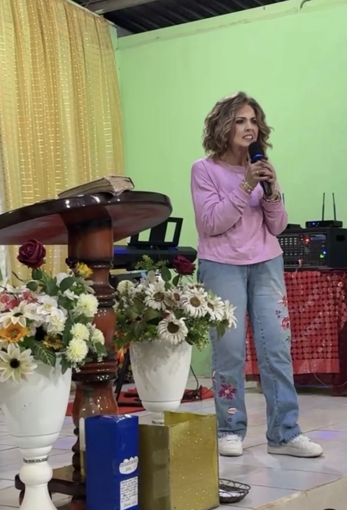 A woman with curly hair wearing a pink top and jeans holding a microphone on a stage decorated with flowers.