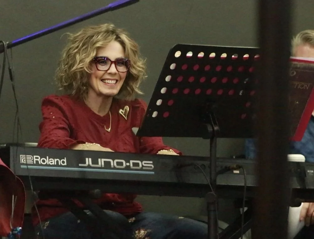 A woman with curly blonde hair and red glasses playing a Roland JUNO-DS keyboard, smiling at a music stand with sheet music in front of her.