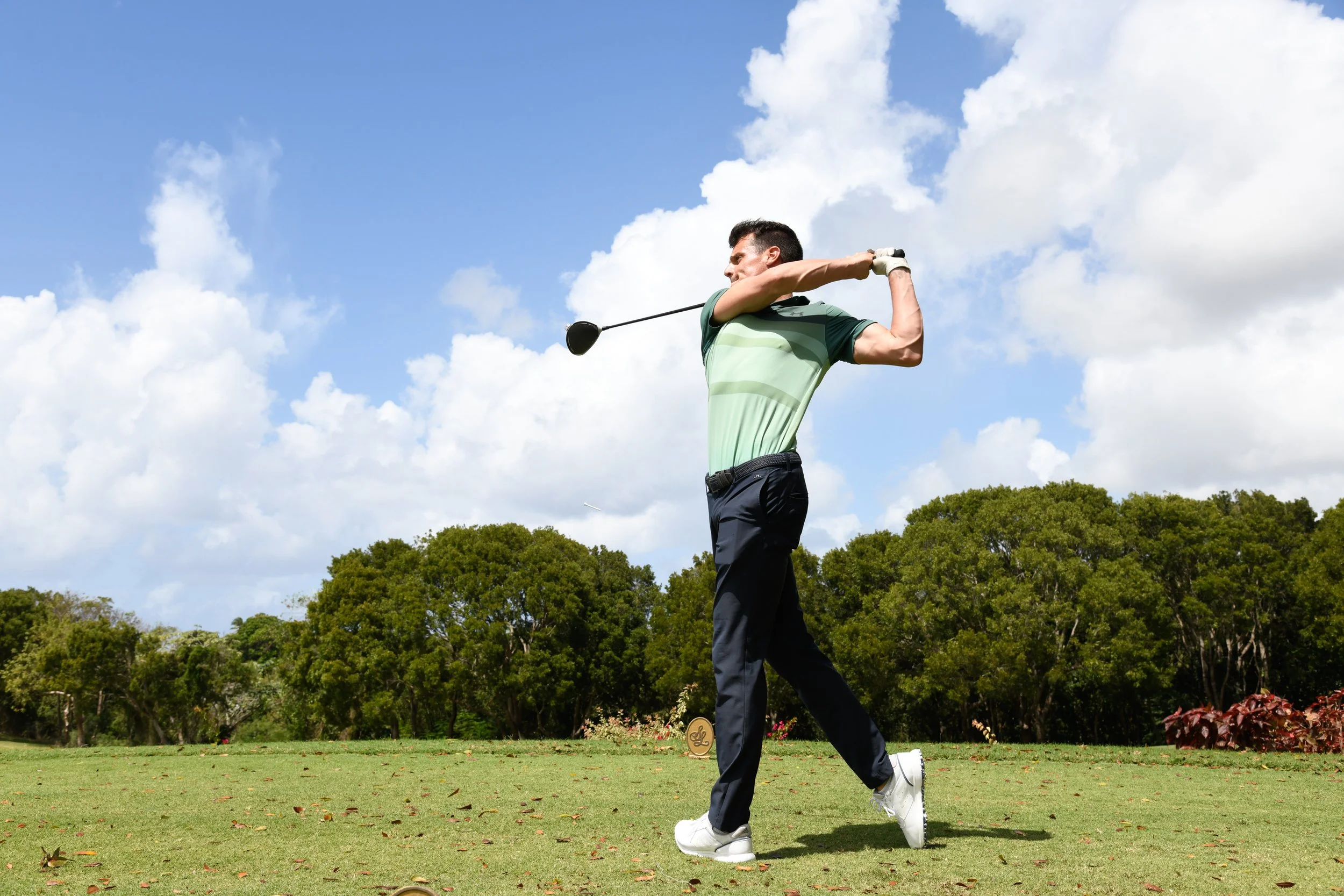 A man swinging a golf club on a golf course with trees and a cloudy sky in the background.