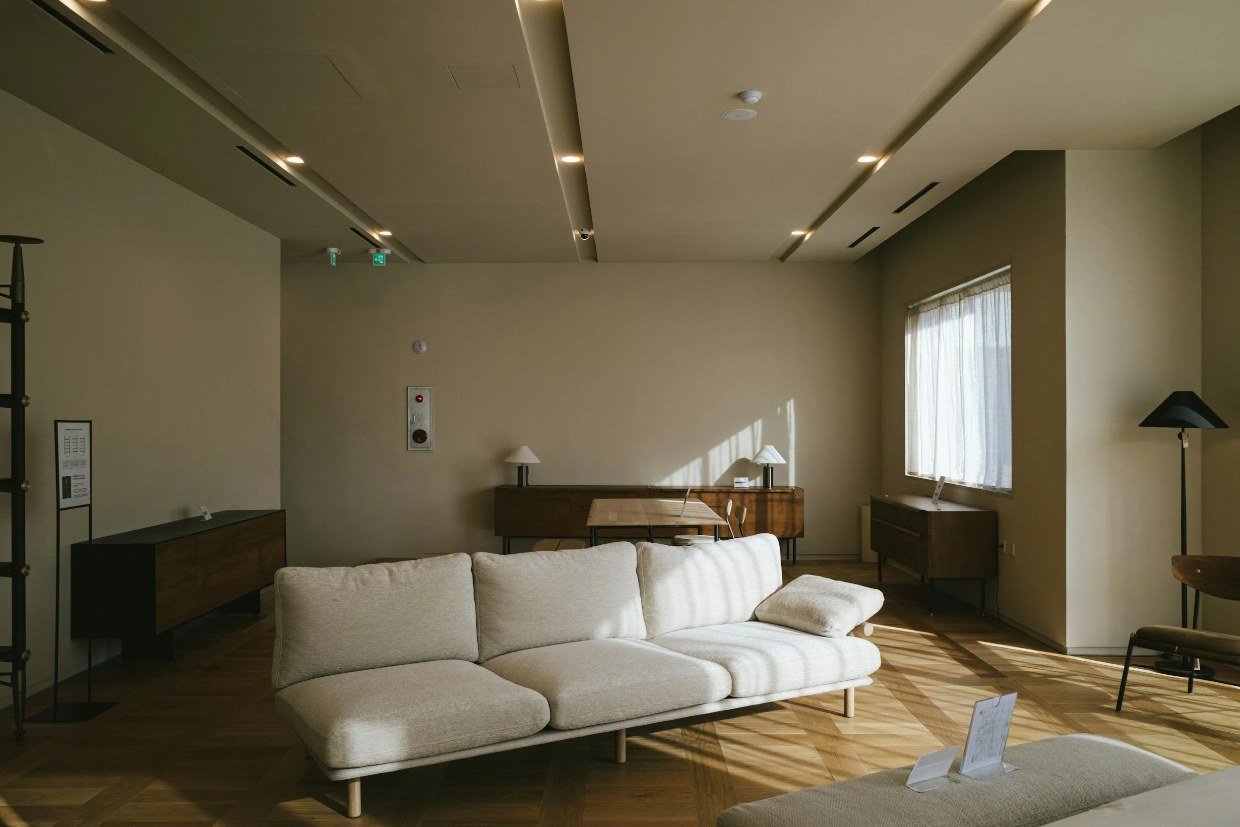 Minimalist living room with beige sofa, wooden floor, sunlight through a window, and modern furniture against cream-colored walls.