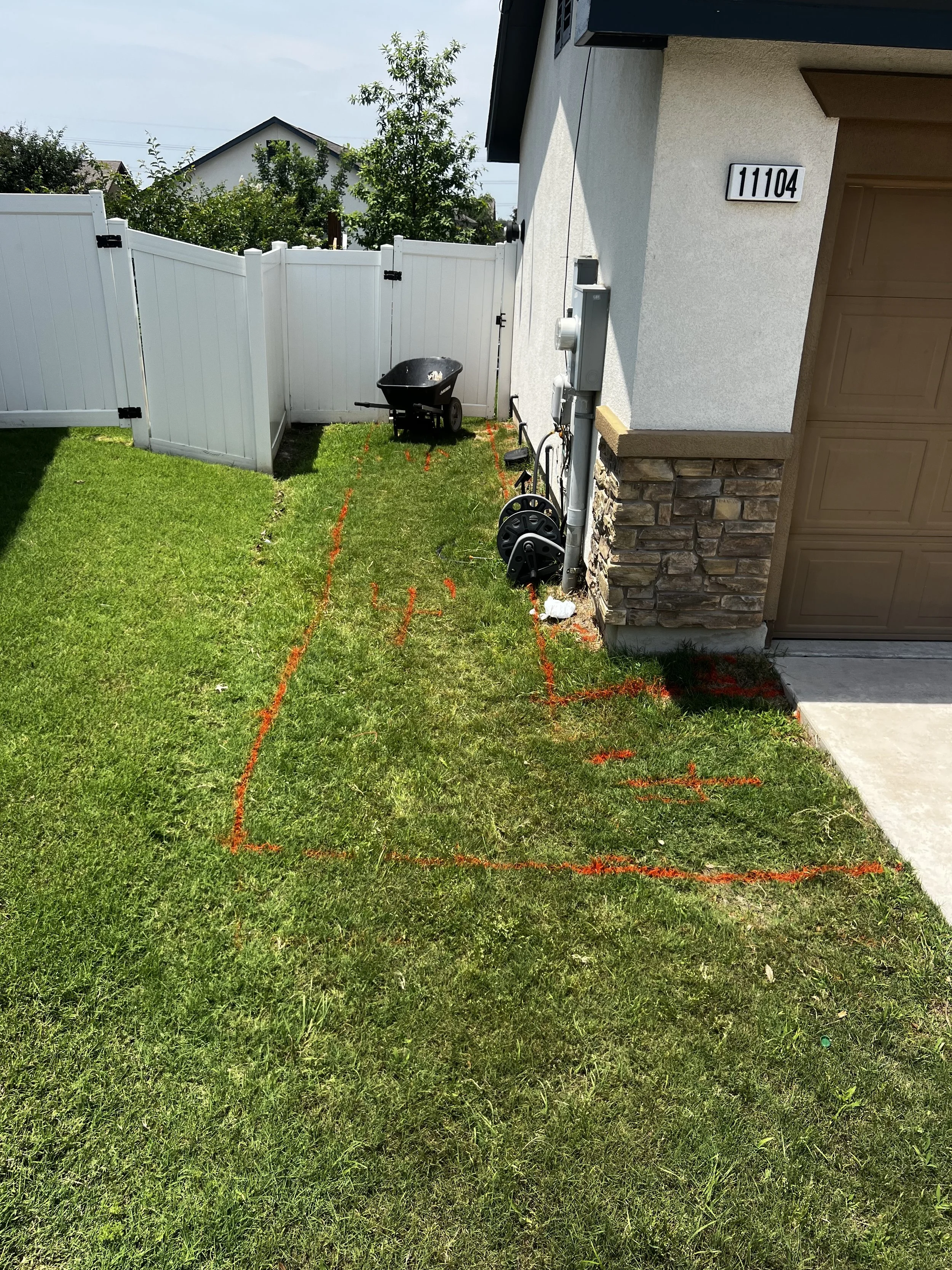 A residential front yard with green grass, orange spray paint markings, a white vinyl fence, a gate, a wheelbarrow, and the house number 11104 on the wall. There are power lines and trees in the background.